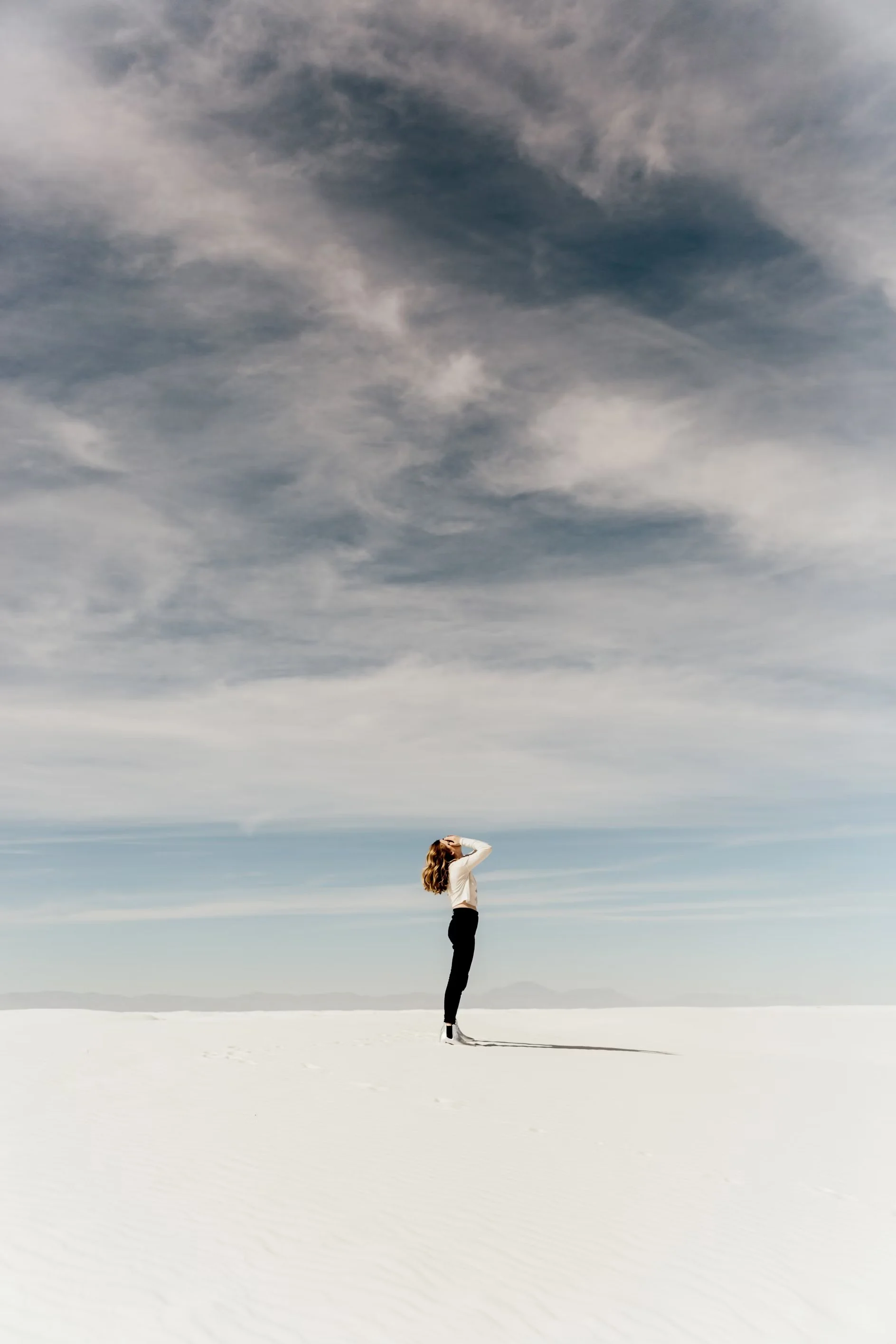 A woman standing alone in a vast, white, sandy desert under a cloudy sky, looking upwards with her hand shielding her eyes.