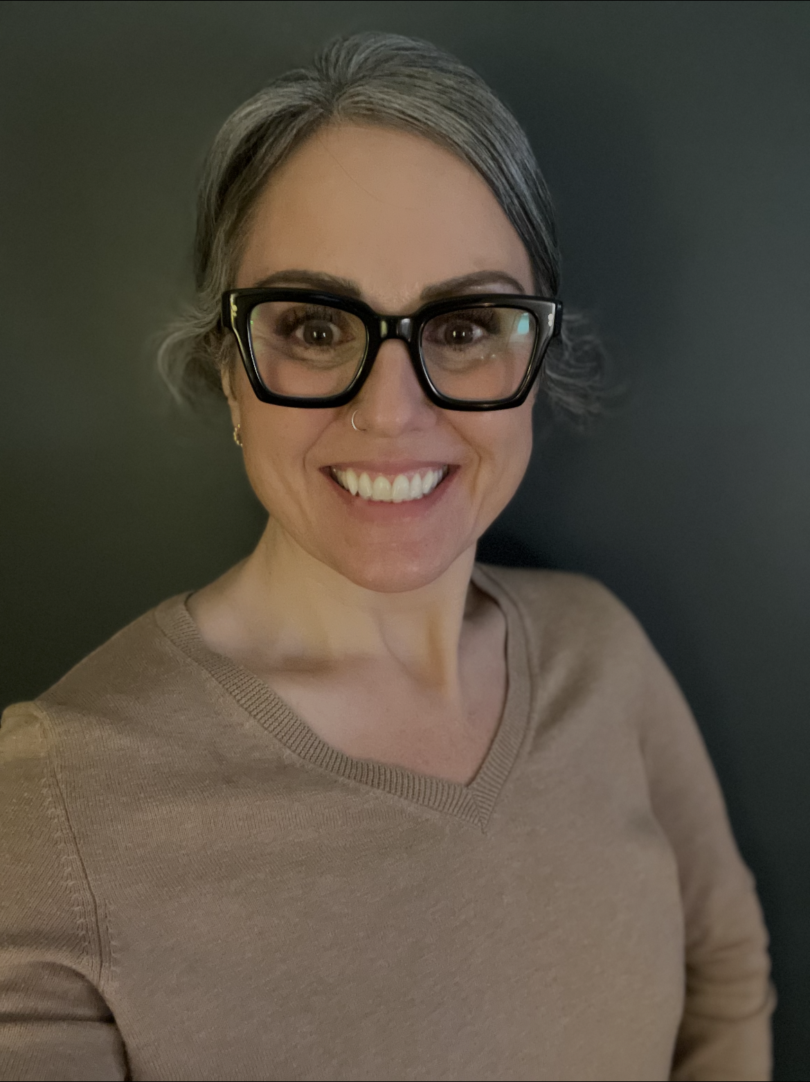 A woman with short gray hair, wearing large black glasses, a small nose ring, earrings, and beige sweater, smiling against a dark background.