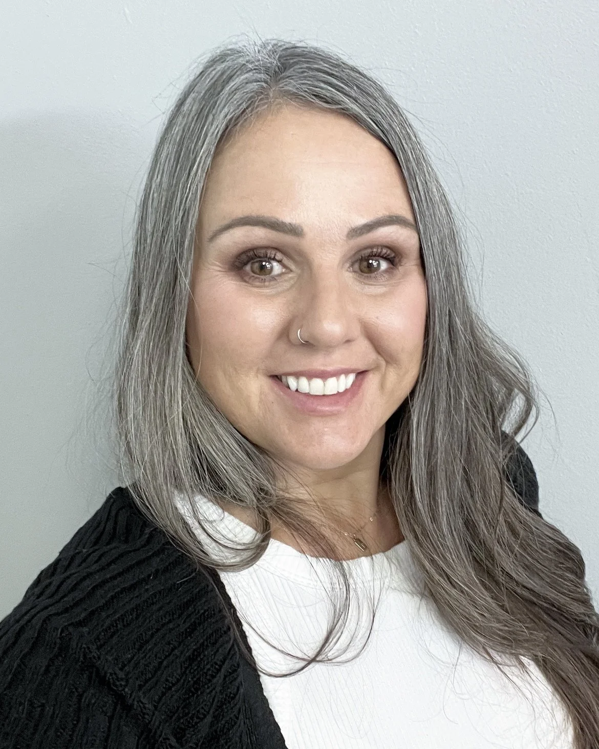 A woman with long gray hair, smiling, wearing a white top and black sweater, and a small nose piercing, against a plain white wall.