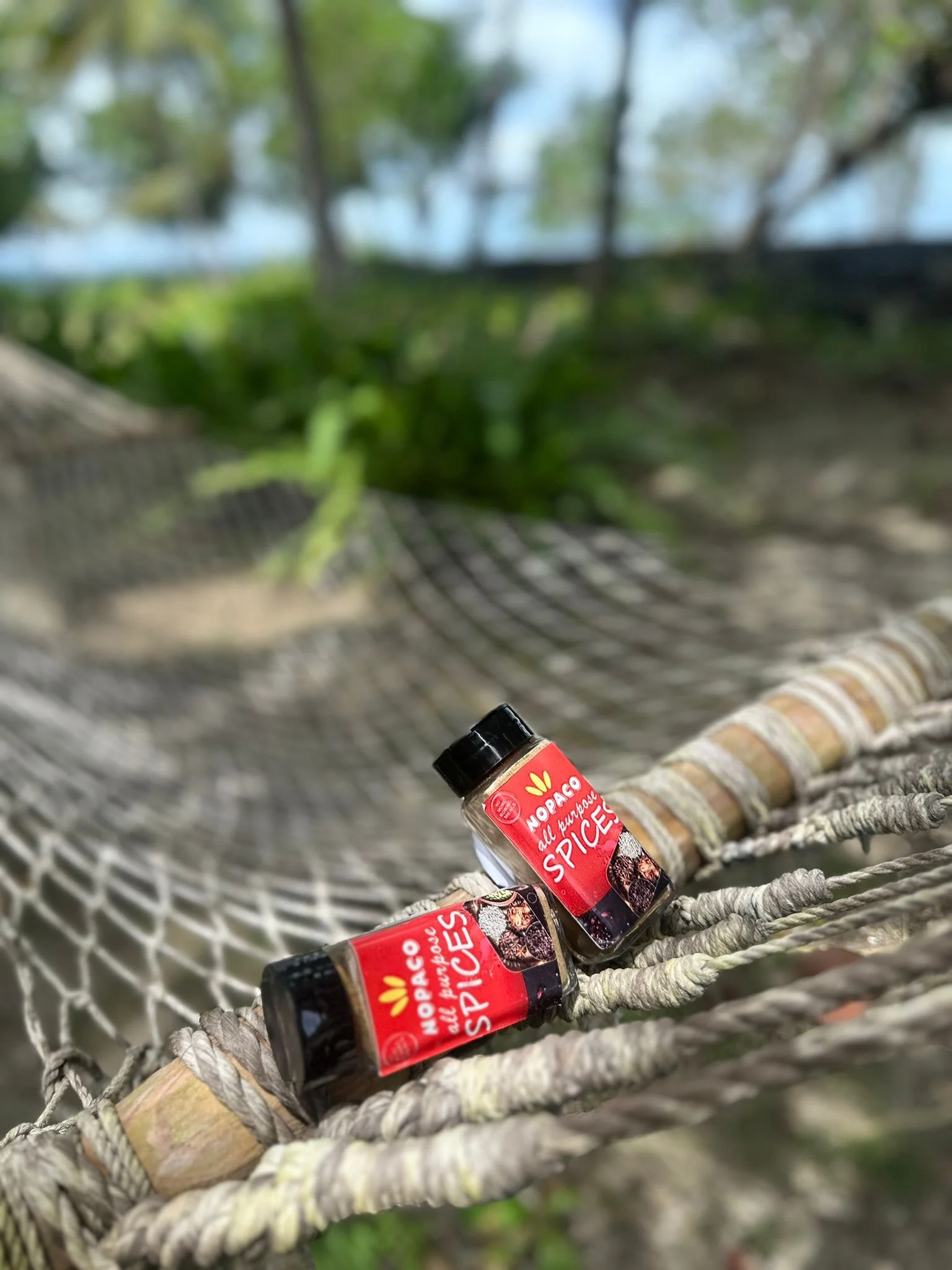 Two bottles of NOPACO All Purpose Spices resting on a woven hammock outdoors with trees and a lake in the background.