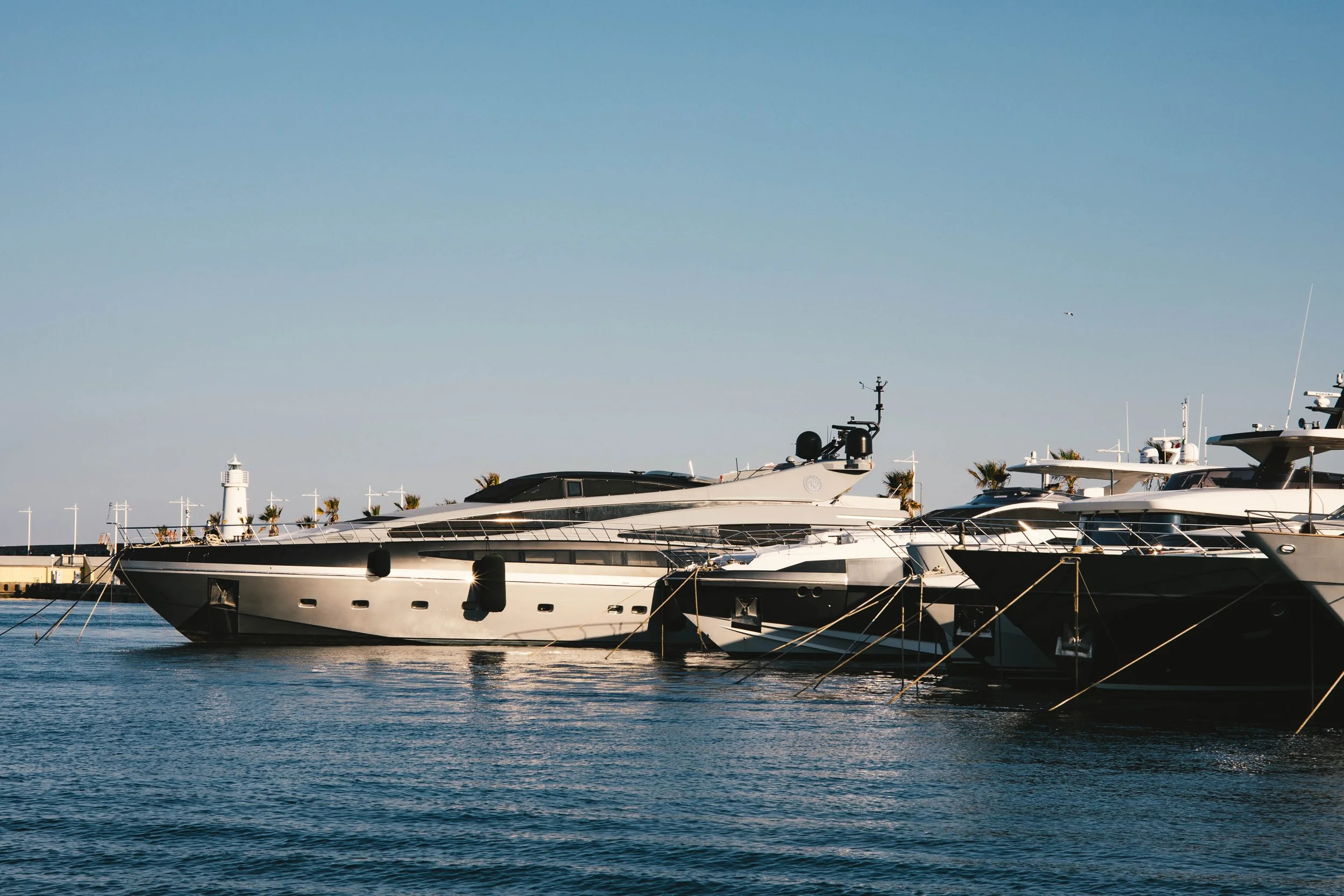 Several luxury yachts docked at a marina with a lighthouse and palm trees in the background under a clear blue sky.