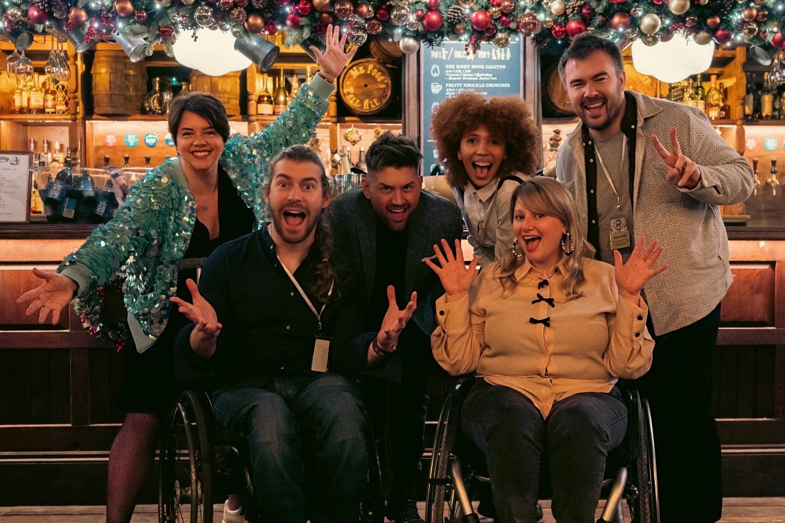 Six smiling friends pose in a festive, Christmas-decorated bar, including two people using wheelchairs.