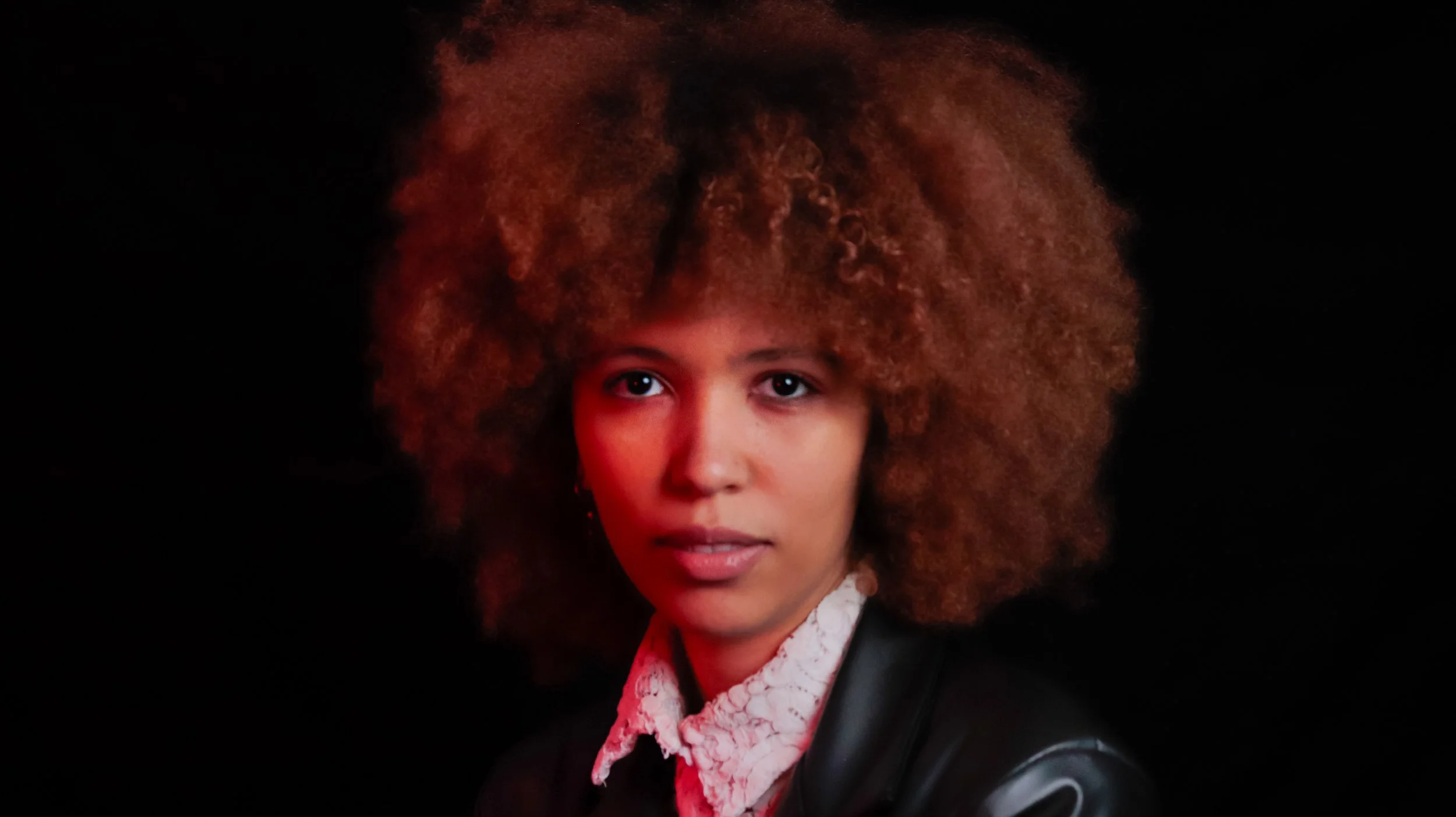 Mixed-race woman with auburn coloured afro wearing a lace shirt and leather blazer. They look straight ahead, half their face is lit in red light