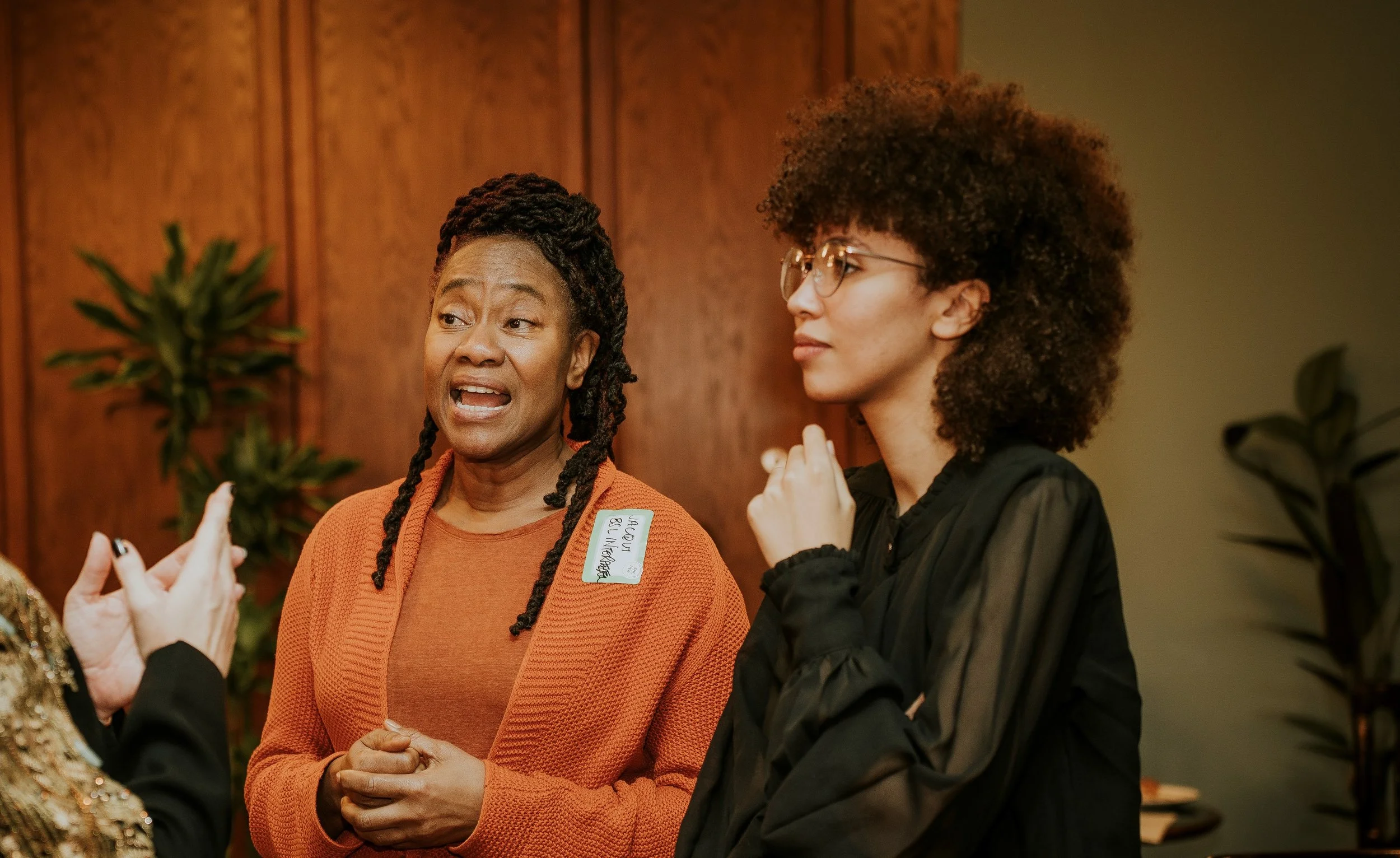 Three women engage in conversation at an indoor gathering, with one woman speaking and wearing a name tag.