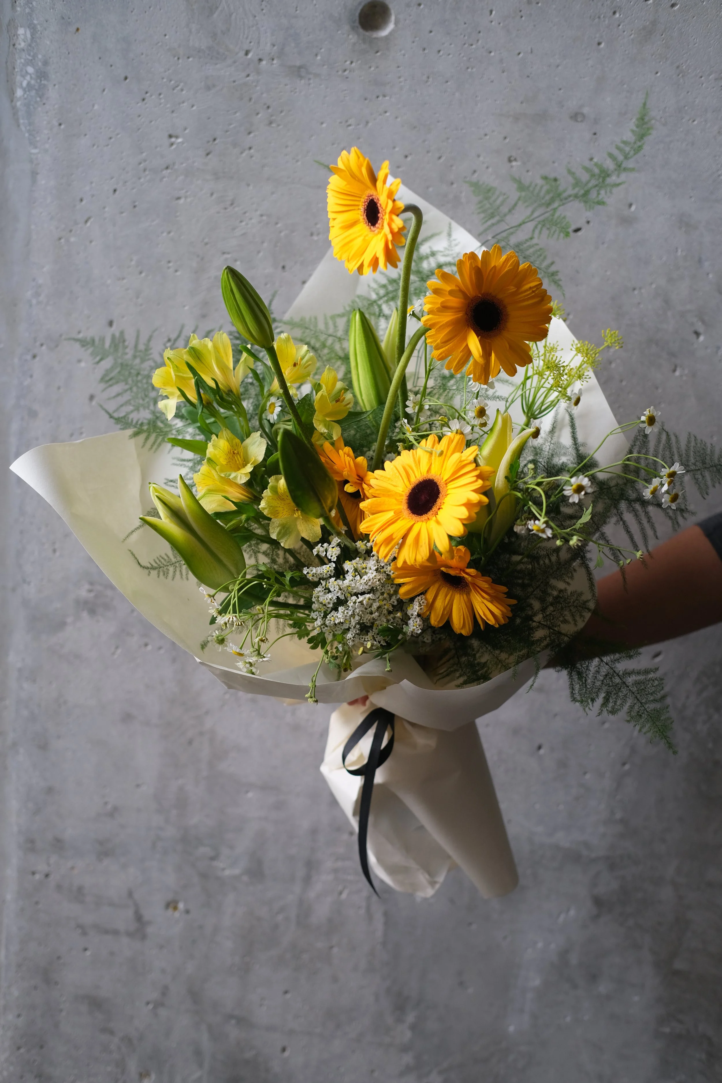 Close-up of Amber Flare Mother's Day bouquet featuring bright yellow gerberas, green lily buds, and chamomile flowers wrapped in minimalist cream paper