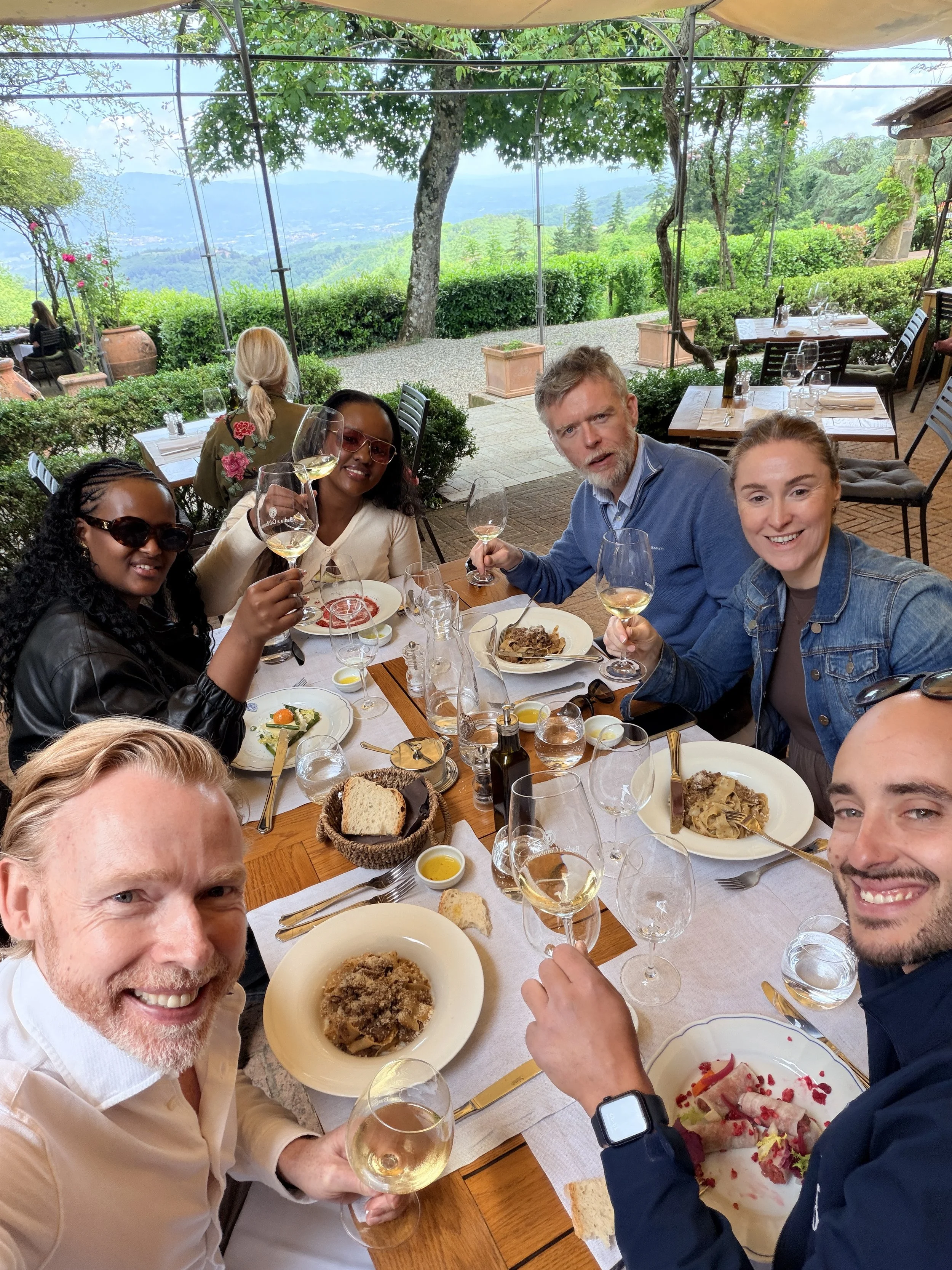 Group of six friends enjoying a meal at the outdoor restaurant of Badia a Coltibuono in Chianti Classico with scenic mountain view, smiling and raising glasses of white wine.