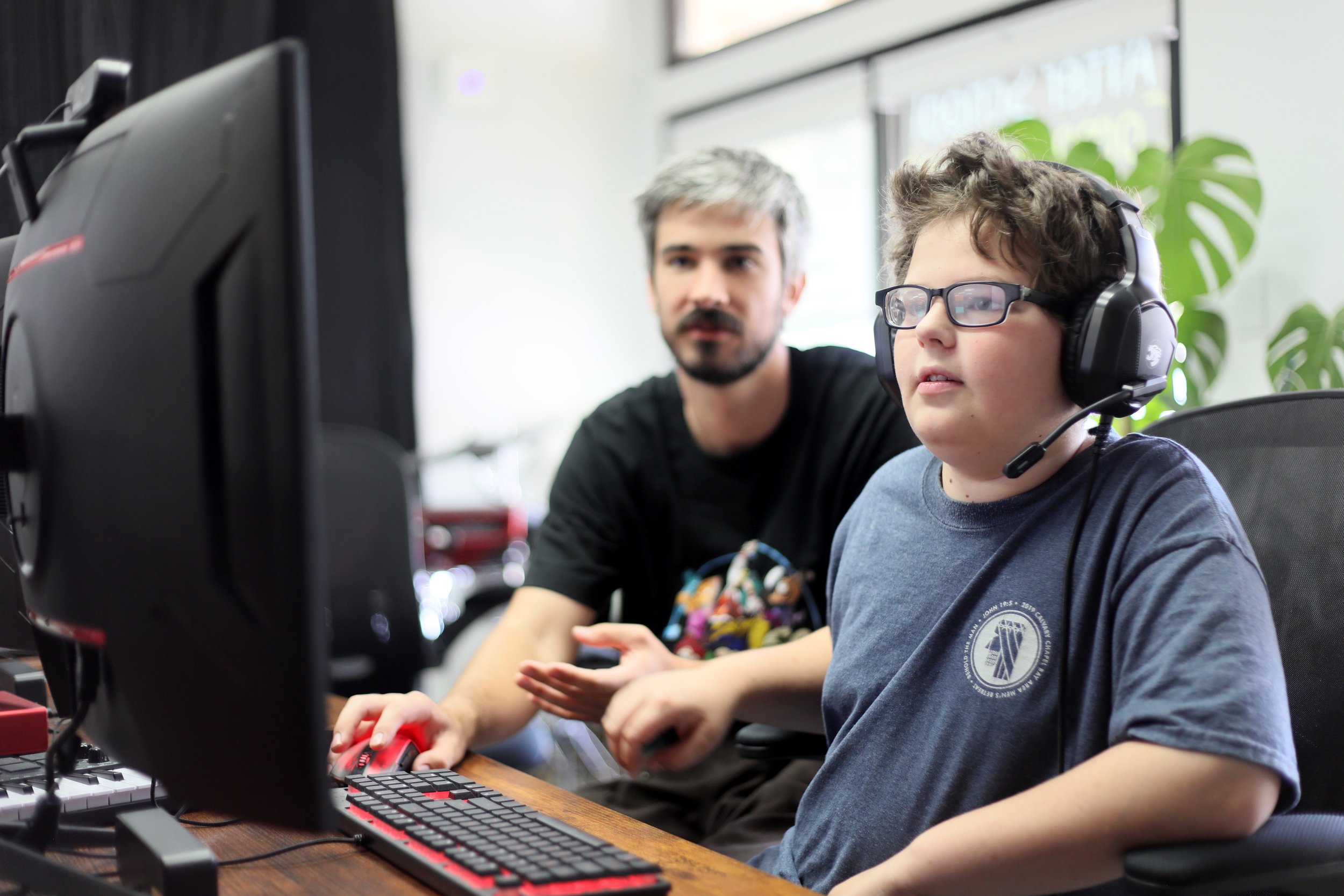 A young boy with glasses and a headset working on a computer with an adult man assisting him, in a bright room with large windows and a green houseplant in the background.