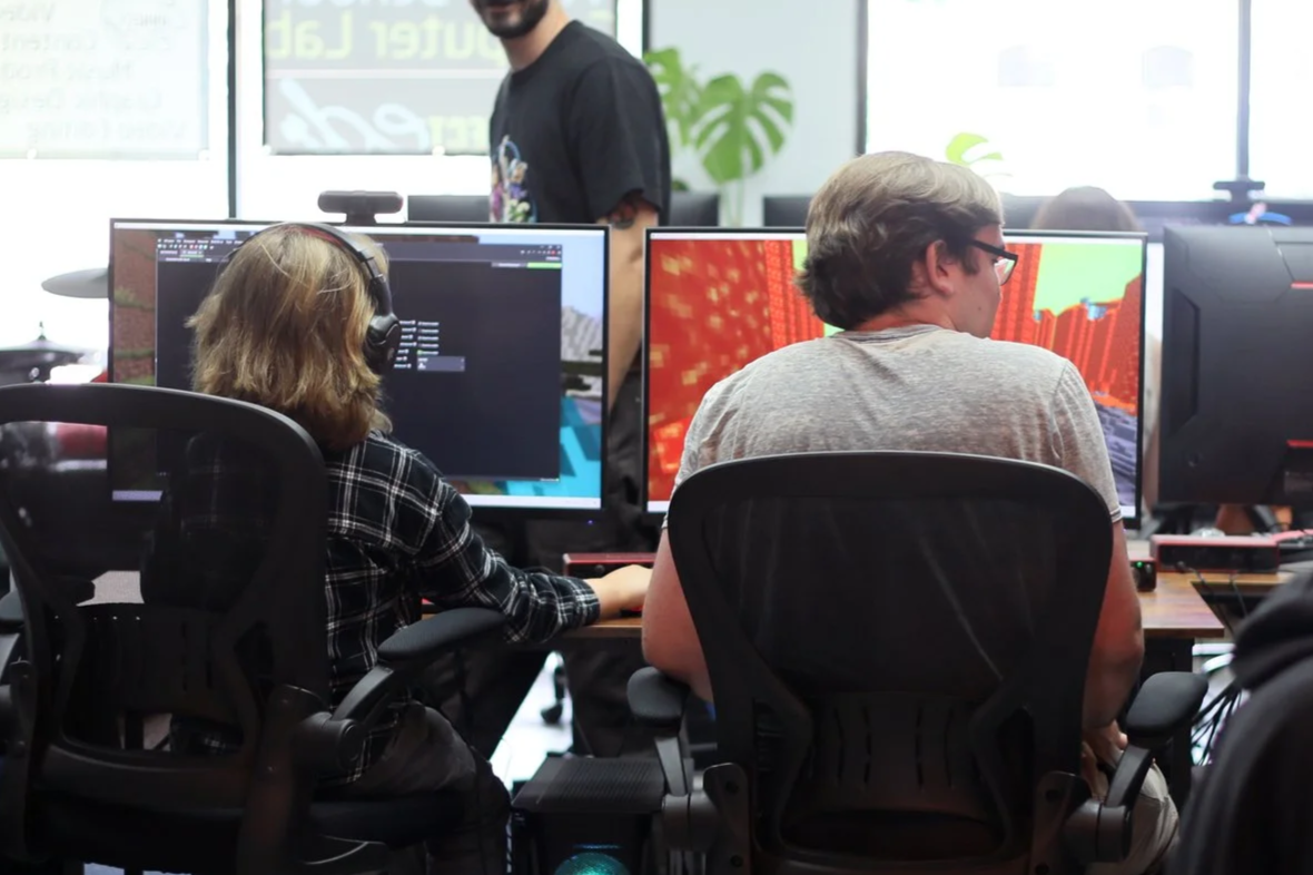 Two people seated at desks with computer monitors, one with headphones and the other with glasses, in an office environment with large windows and plants, while a person stands behind them.