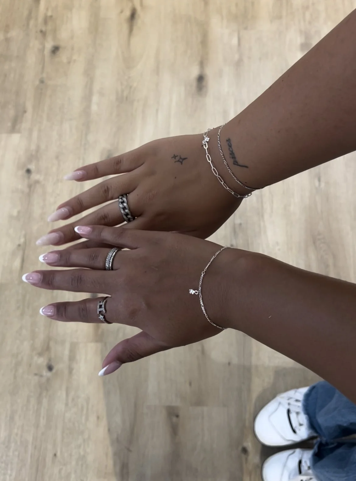 Two hands with rings and bracelets, displaying tattoos on wrists, against a wooden floor background.