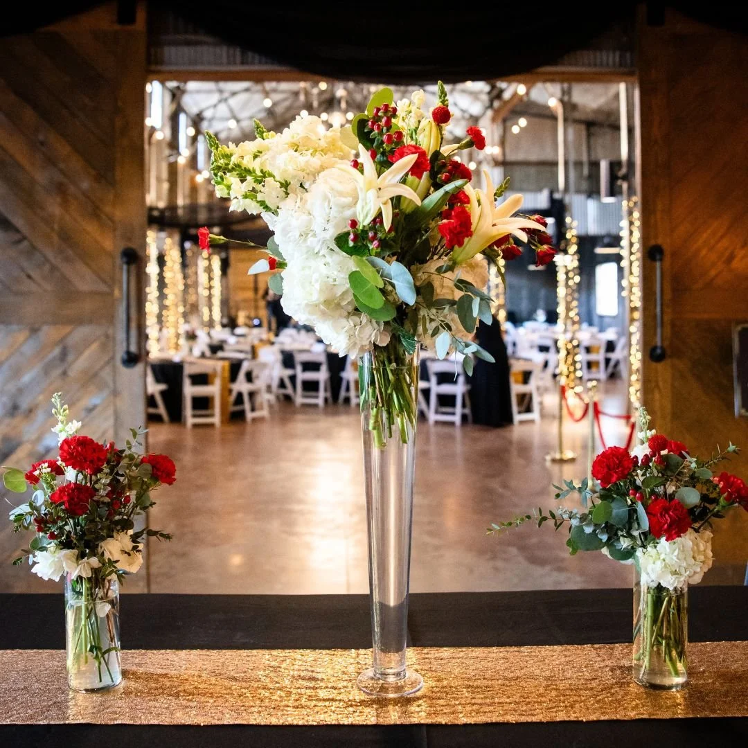 Elegant floral arrangement with white lilies and hydrangeas, and red accents in tall and small vases on a table, set in a rustic wooden venue decorated with string lights.
