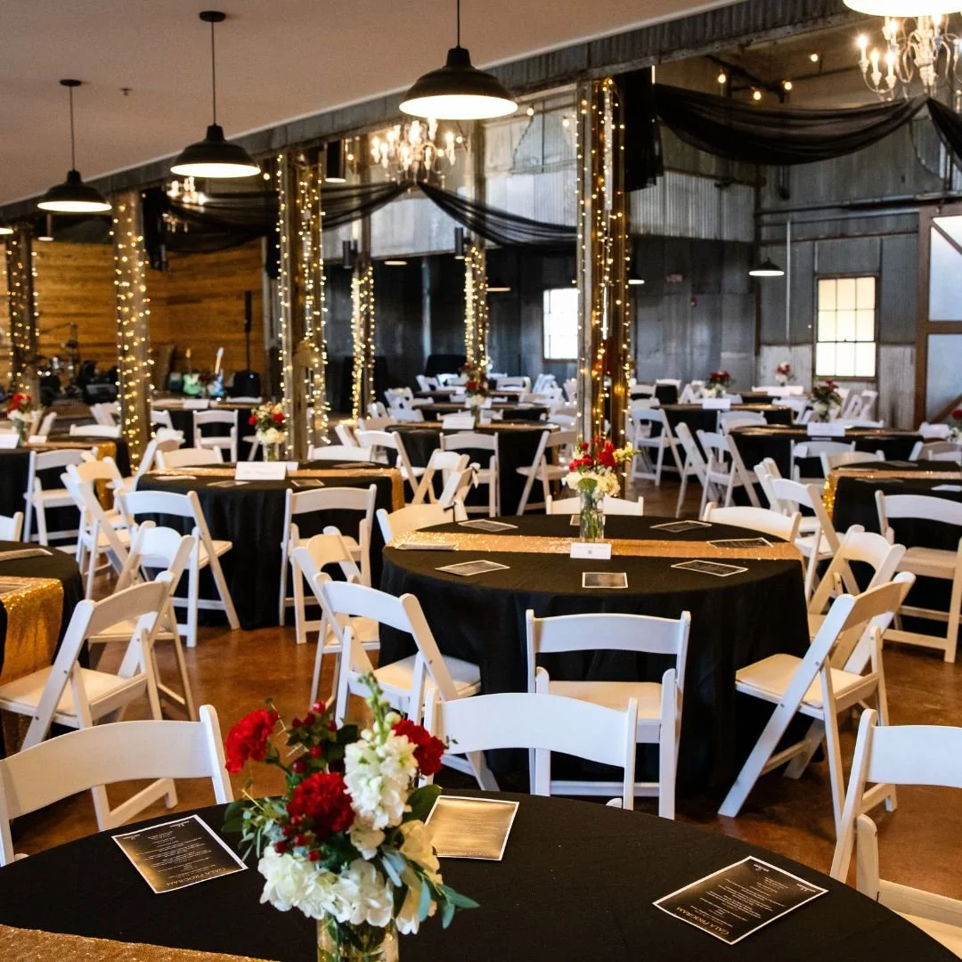 Elegant event space decorated with black tablecloths, white chairs, floral centerpieces, string lights, draped black fabric, and chandeliers.
