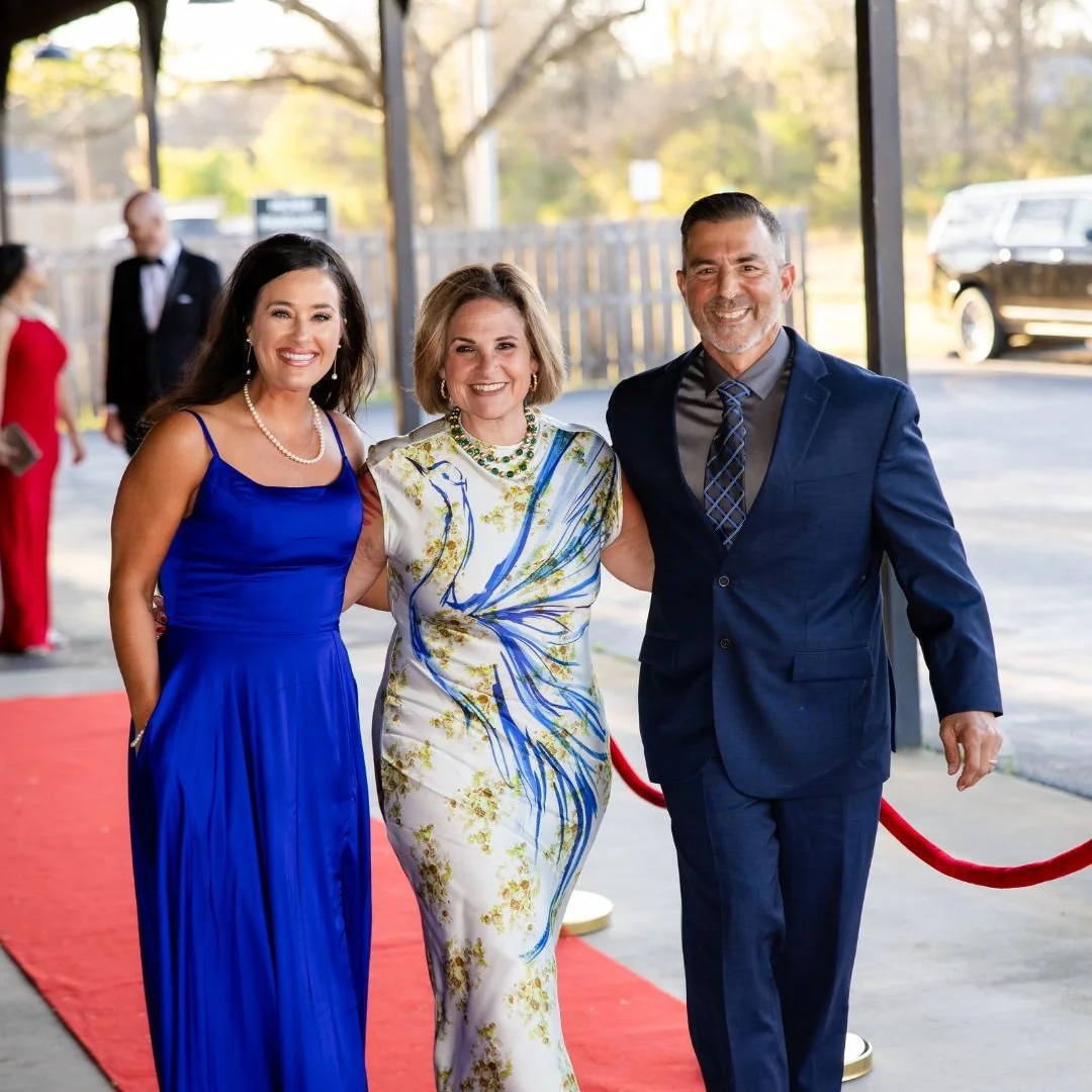 Three people dressed in formal attire standing on a red carpet outside, smiling at the camera. There are other guests and a black vehicle in the background.