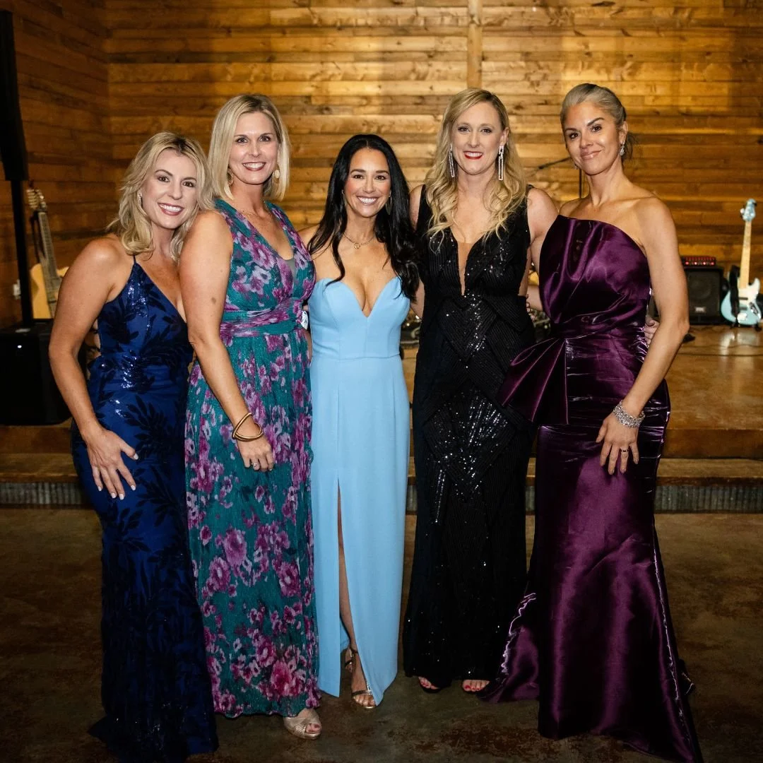 Five women in formal dresses standing together indoors against wooden wall, smiling at the camera, with guitars and music equipment in the background.
