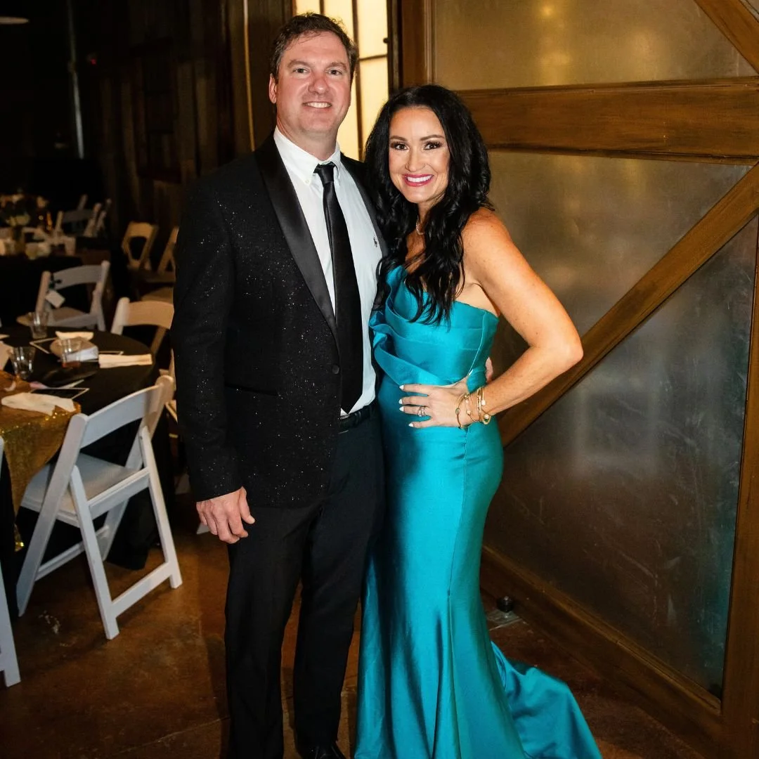 A man in a black suit with a white shirt and tie beside a woman in a strapless turquoise gown, smiling for a photo at an indoor event.
