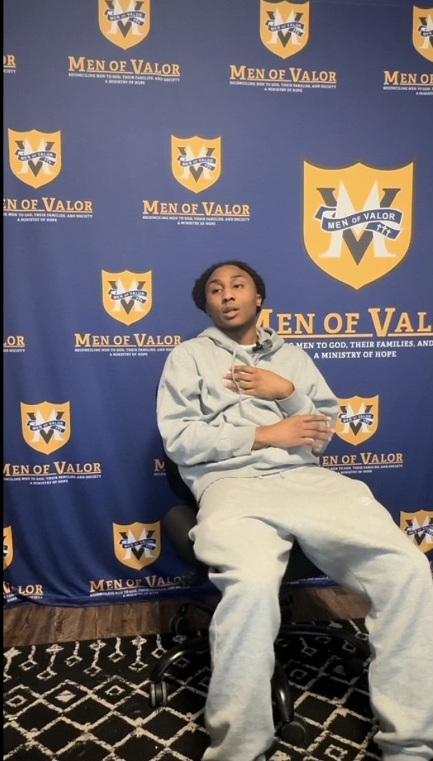 A young man in a grey hoodie and sweatpants sitting in a wheelchair in front of a blue backdrop with the Men of Valor logo and text.