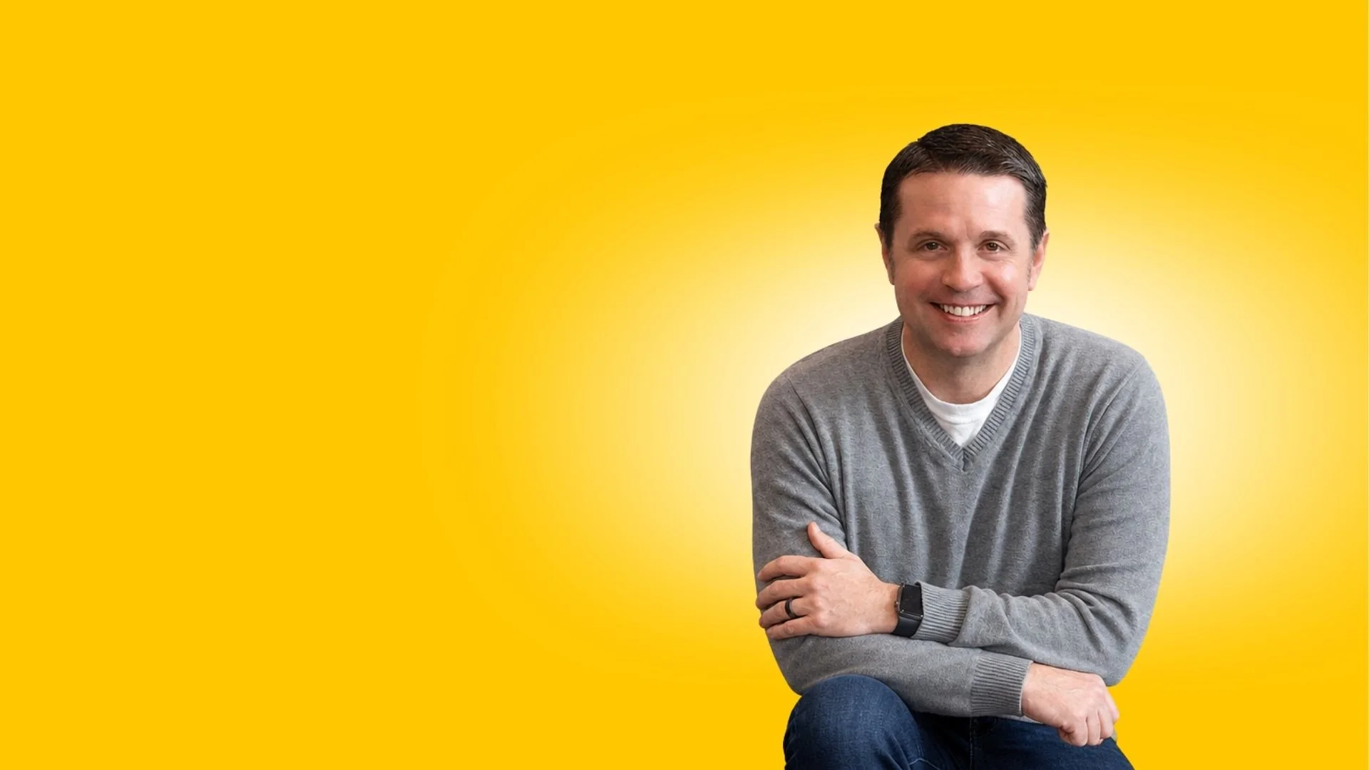 Smiling man sitting with arms crossed in front of a yellow background