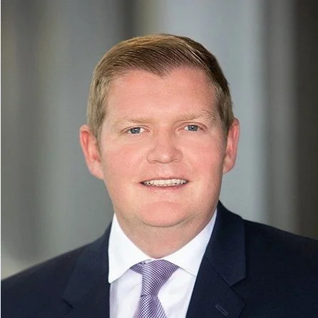 Smiling Ryan Carlson, Esq, Board Chairperson, in business attire with a suit and tie, neutral background