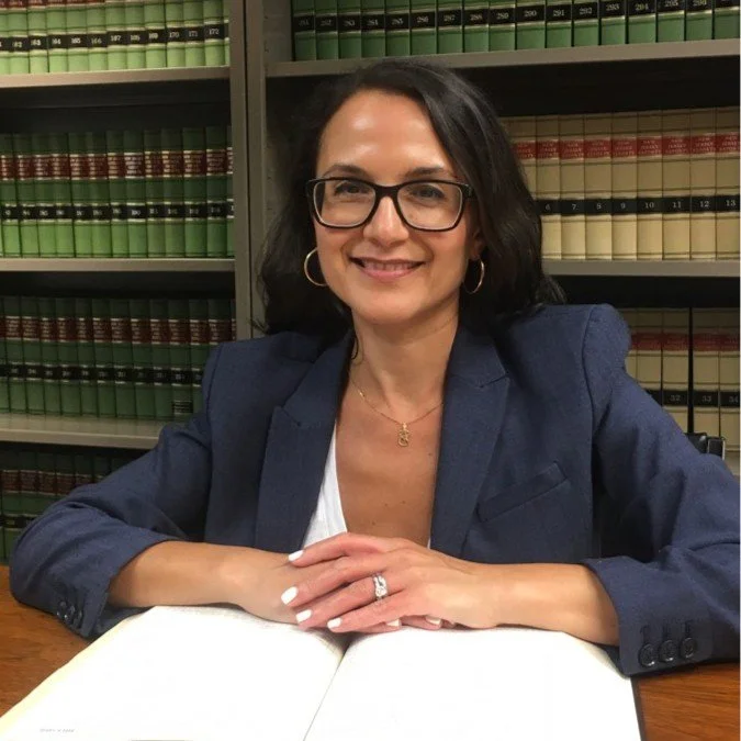 Erika Kerber, Esq., President/Executive Director, in a blue blazer sits at a desk with open books, surrounded by shelves full of legal books.