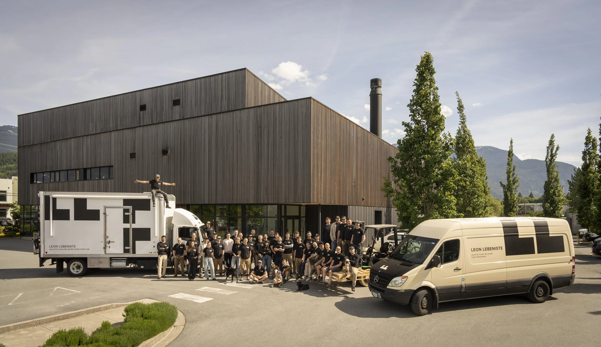 Group of people standing in front of a modern wooden building with trucks and vans nearby, and mountains in the background.