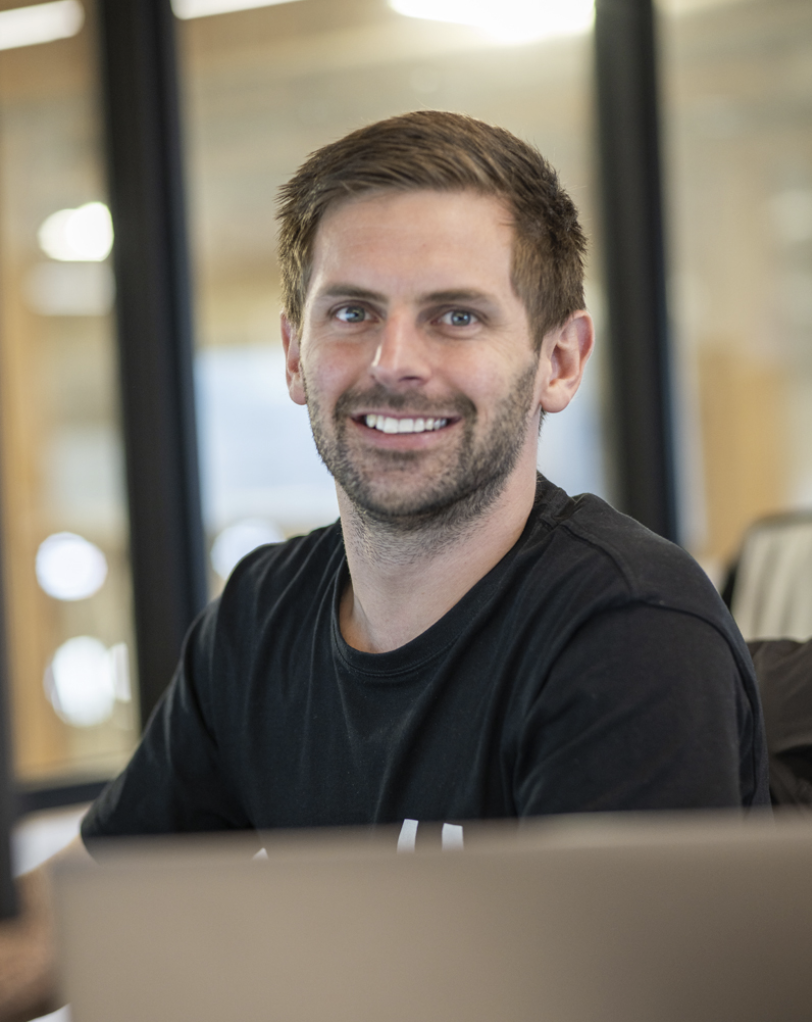 A smiling man with short brown hair and a beard, wearing a black T-shirt, sitting in an office environment with large windows.