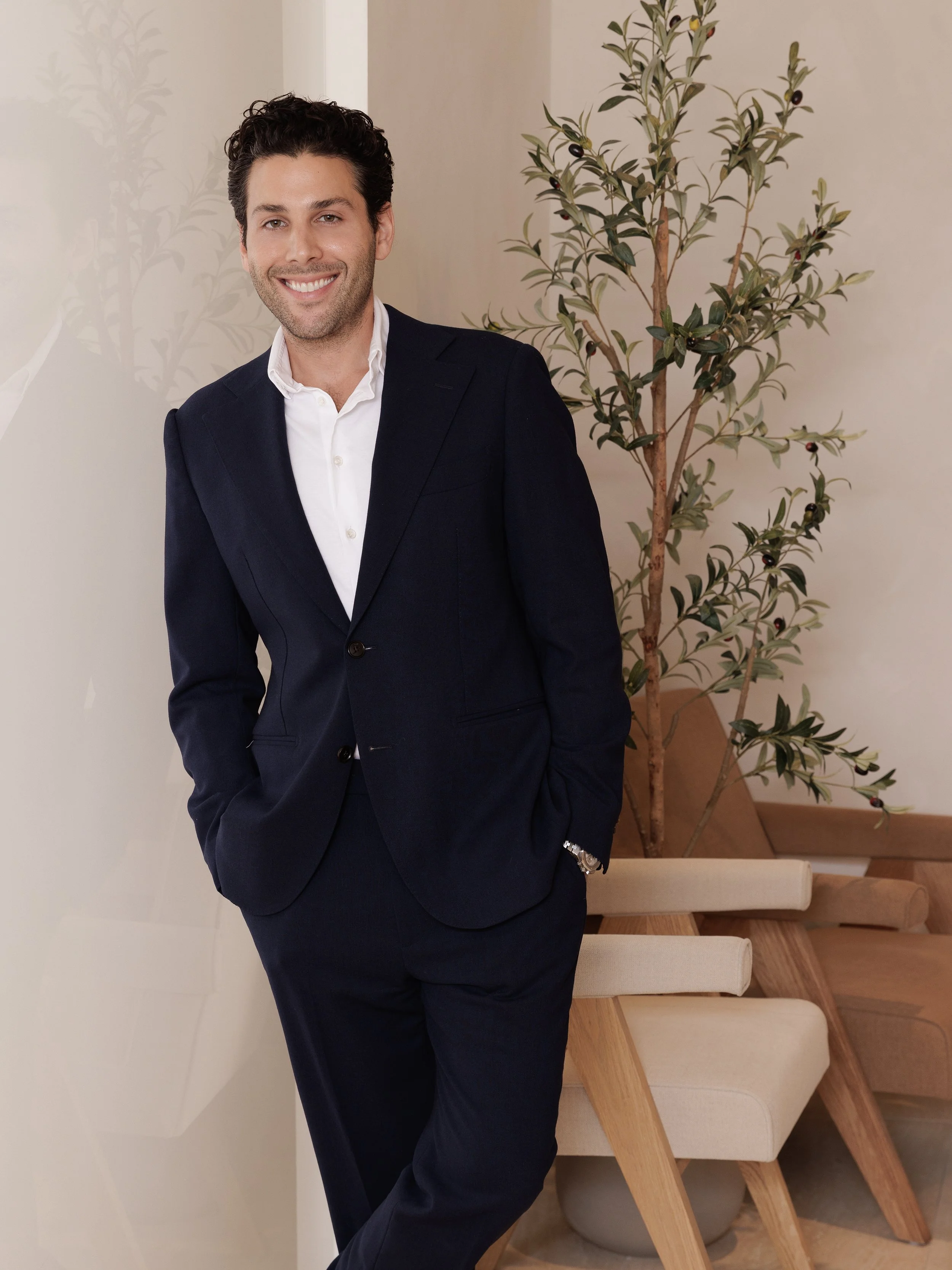 Environmental portrait of a medical professional photographed in his practice's reception area. The relaxed pose, genuine smile, and soft natural light convey approachability and trust, while the modern interior — warm beige tones, natural wood seati