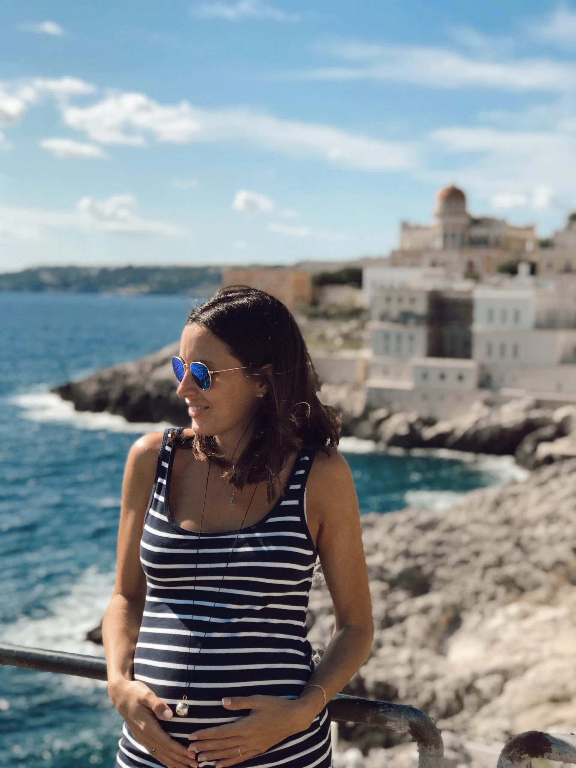 Young pregnant woman in blue and white striped dress, wearing blue Rayban sunglasses, standing by the sea in Italy with a white castle in the background, a blue sky with a few clouds.