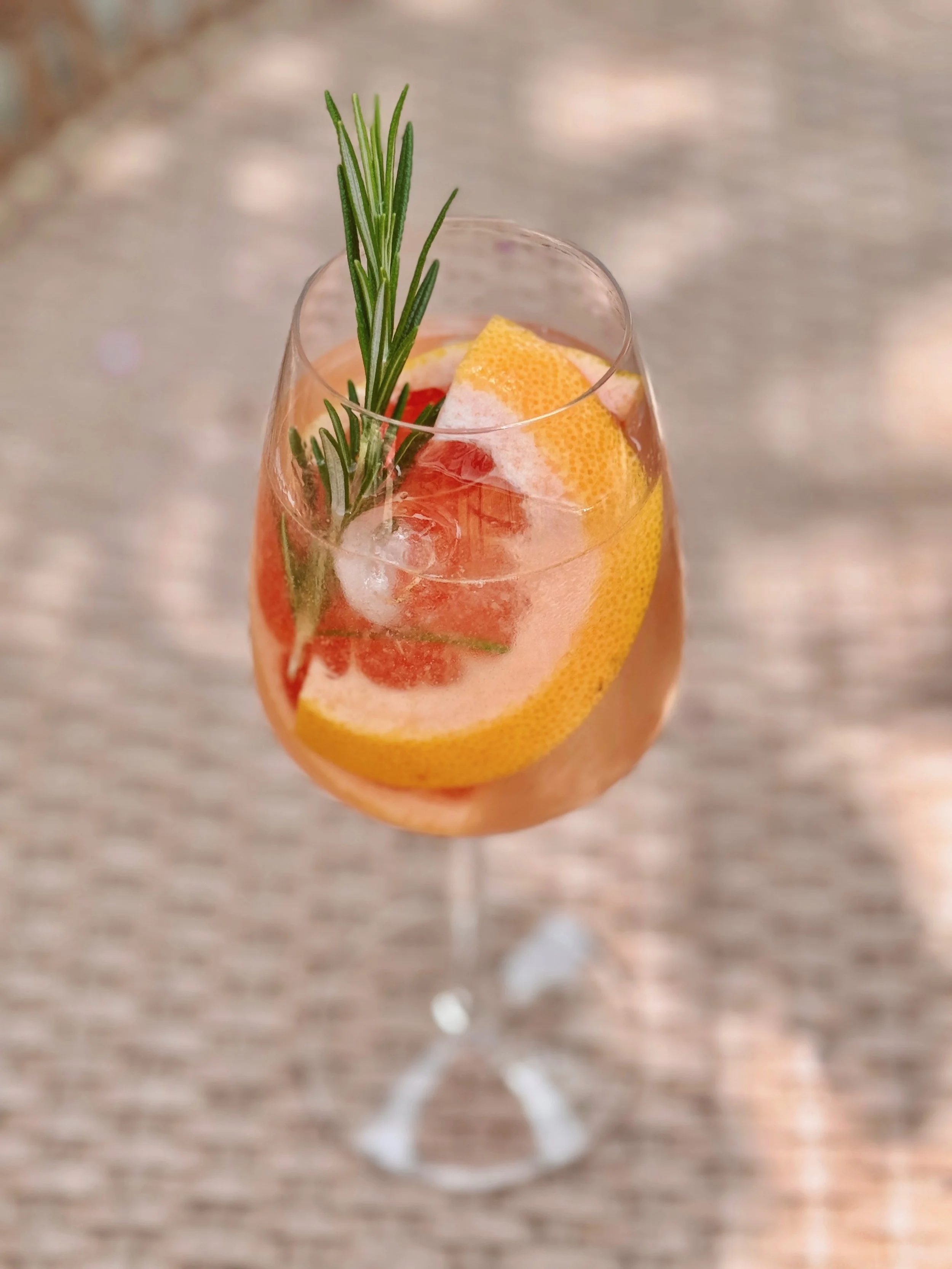 Un verre à vin rempli d'une boisson rose avec une tranche de pamplemousse, une branche de romarin, et des glaçons. Recette Spritz sans alcool.