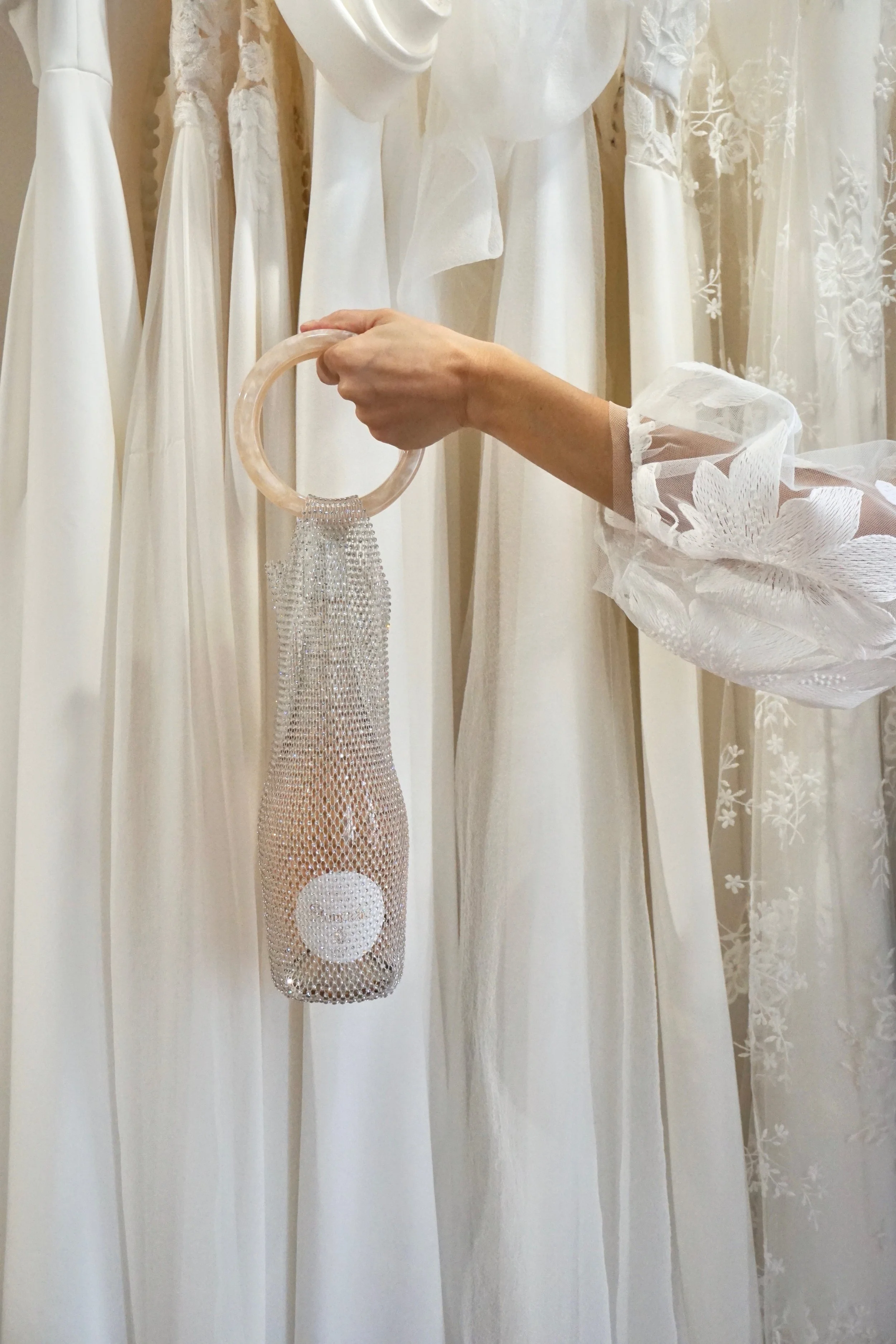 One hand holds a bottle holder with a sparkly design, with a bottle of non-alcoholic rosé wine inside. In the background, several white wedding dresses and lace fabrics hang.