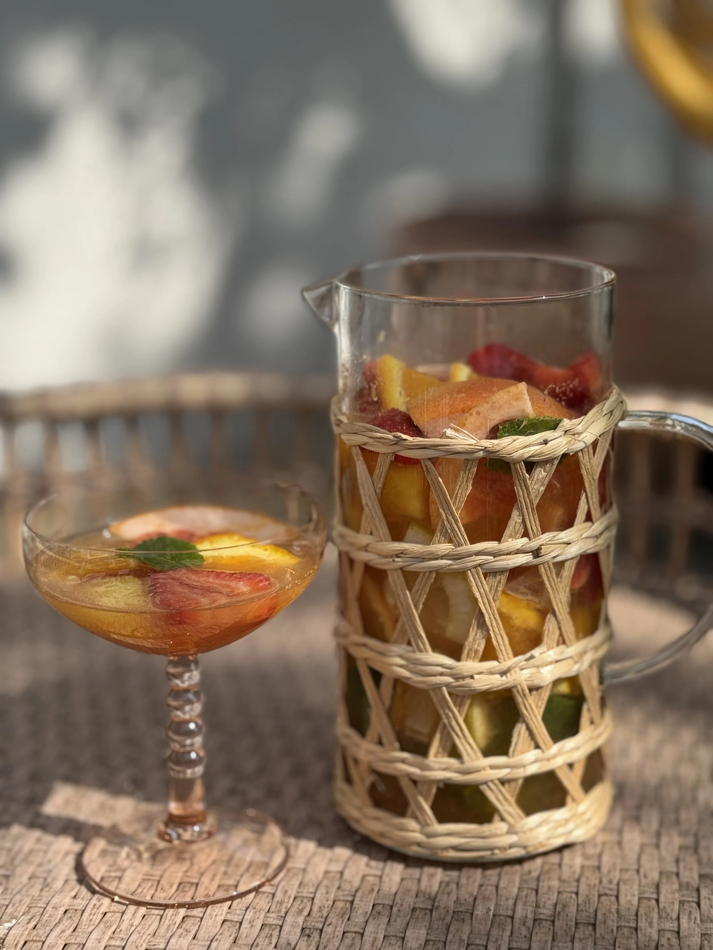 Un verre à cocktail rempli de fruits coupés, avec des feuilles de menthe, placé à côté d'une carafe en verre avec un nœud en raphia, contenant des morceaux de fruits, sur une surface en bois. Recette Sangria rosé sans alcool.