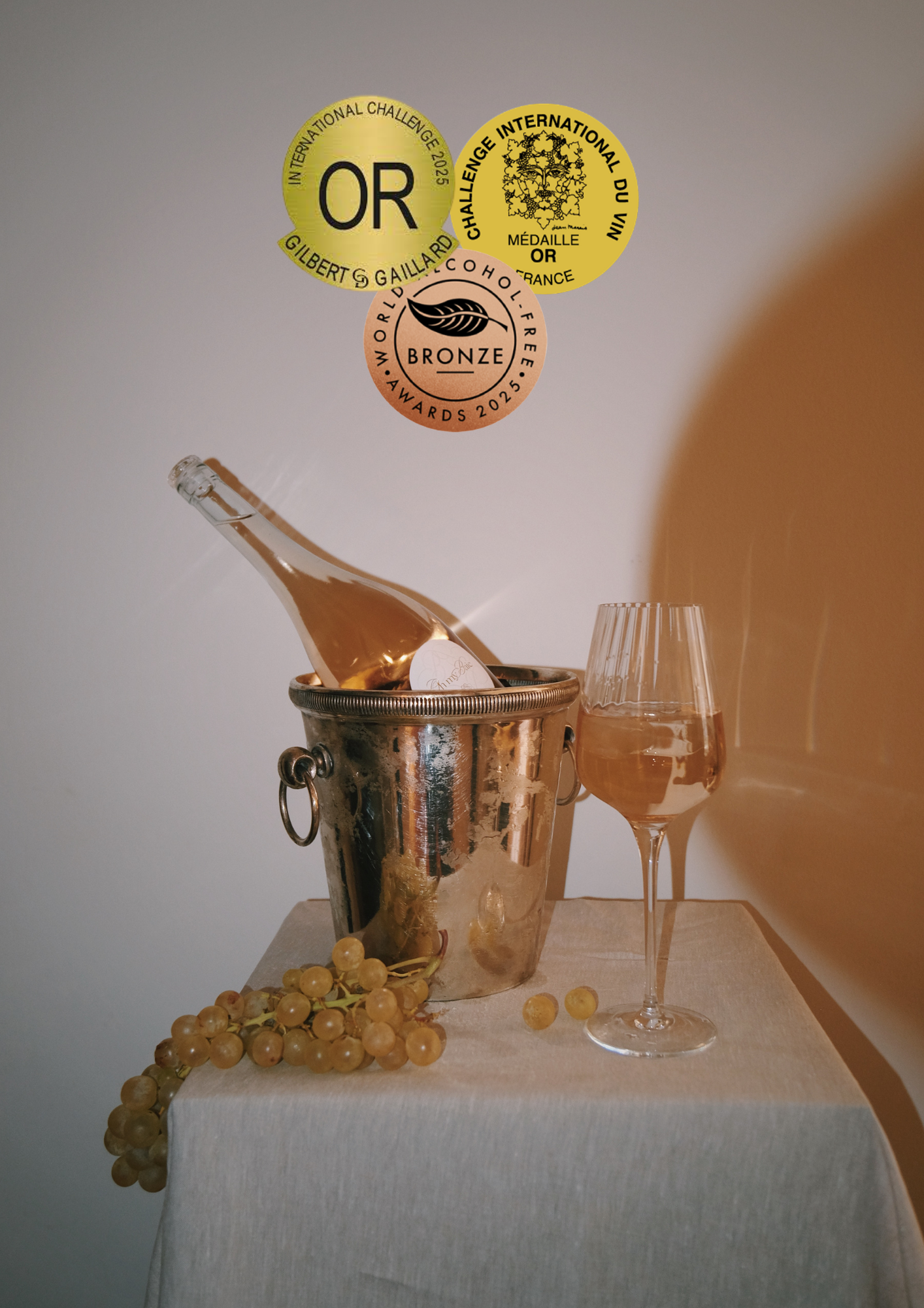 Une bouteille de rosé sans alcool dans un seau à glace, une coupe à vin rosé remplie de vin rosé, un bouquet de raisins blancs, et trois médailles d'or et de bronze au-dessus, sur un fond neutre.