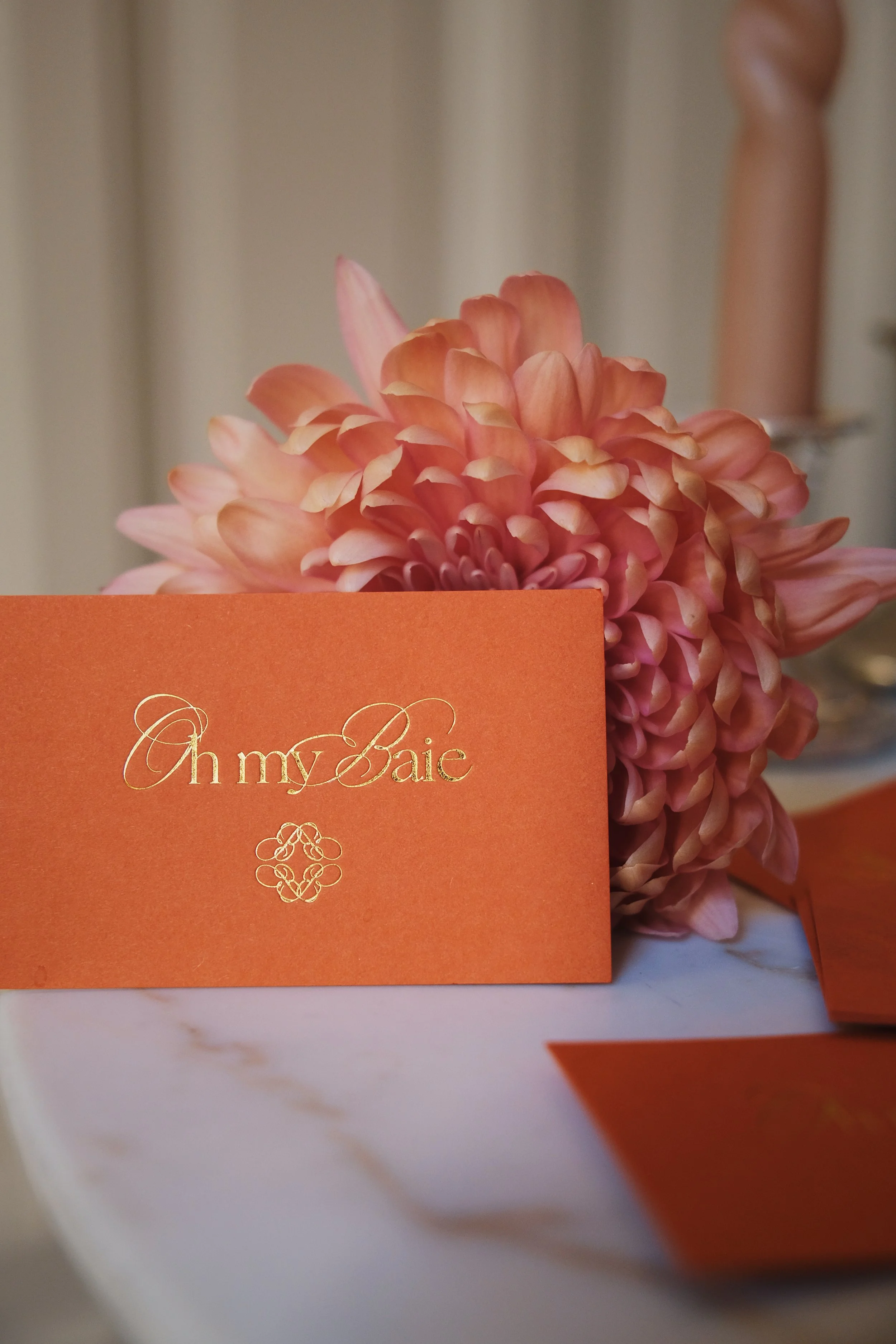 An orange business card with gold Oh my Baie lettering, set on a table next to a bouquet of pink flowers, for a wedding or special occasion.