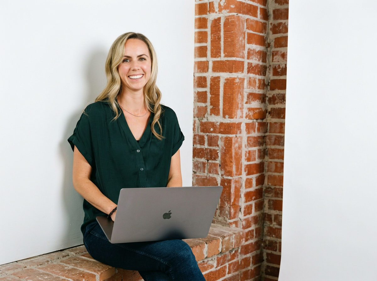 Founder Kristi Jacobsen sitting with her laptop on a brick wall, smiling at the camera.
