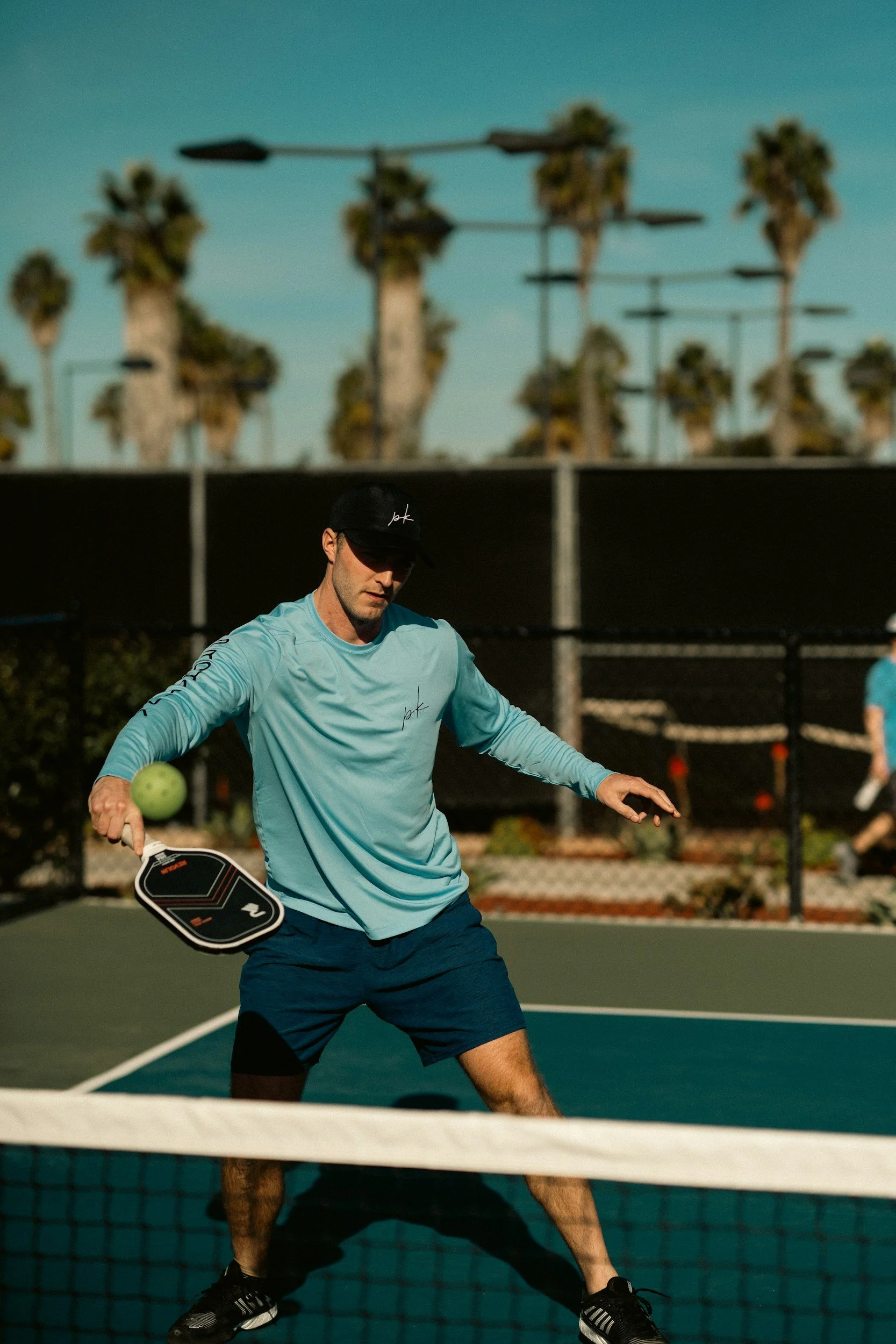 A man wearing a black cap, light blue long-sleeve shirt, and navy shorts is playing pickleball on an outdoor court at dusk. He is holding a paddle in his right hand and has a green pickleball nearby. There are palm trees and a fence in the background.