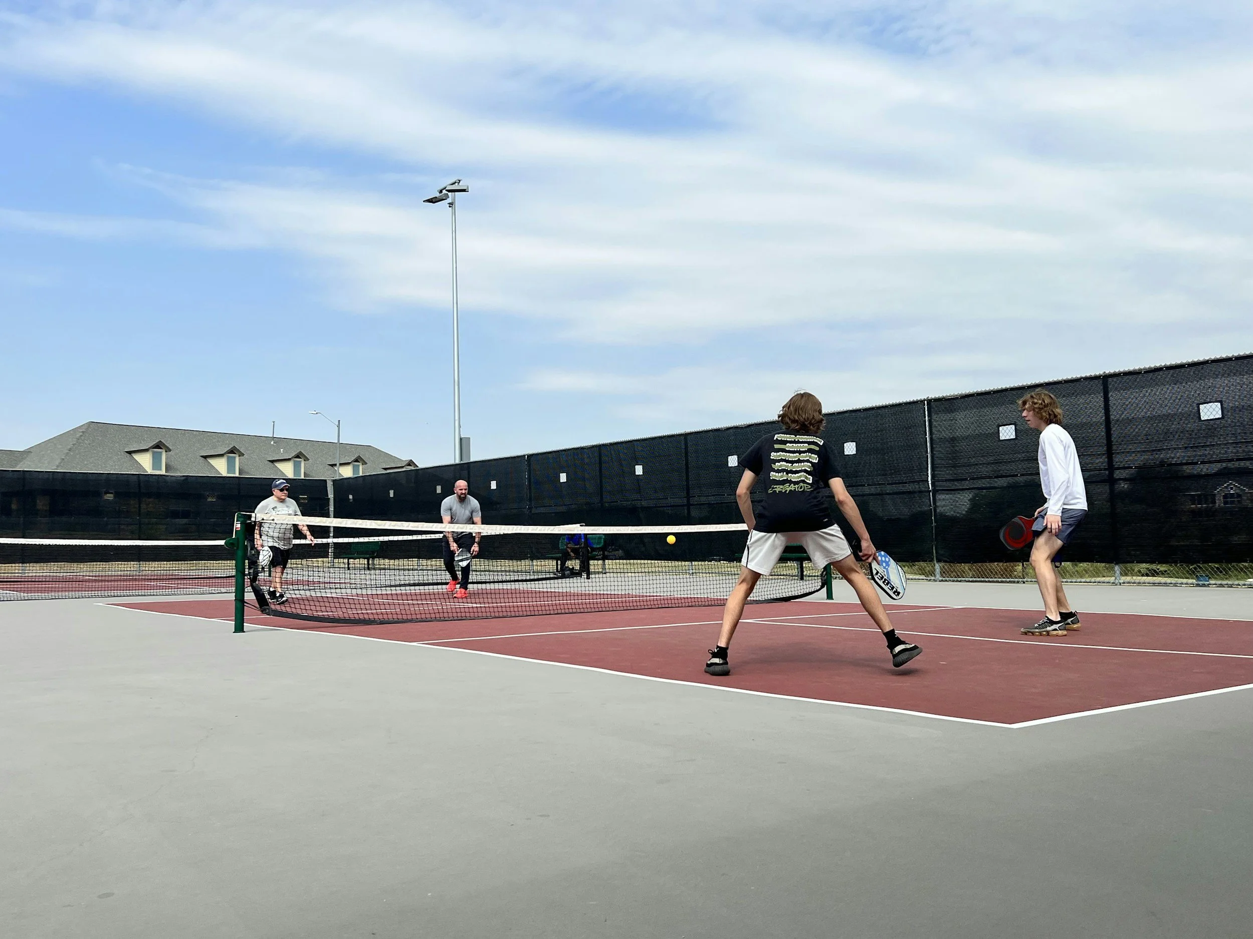 People playing pickleball on an outdoor court with a black windscreen, residential houses in the background, and a partly cloudy sky.