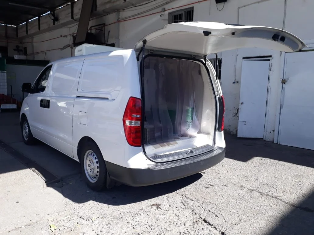 Furgoneta blanca con la puerta trasera levantada, mostrando espacio para carga en un área industrial.