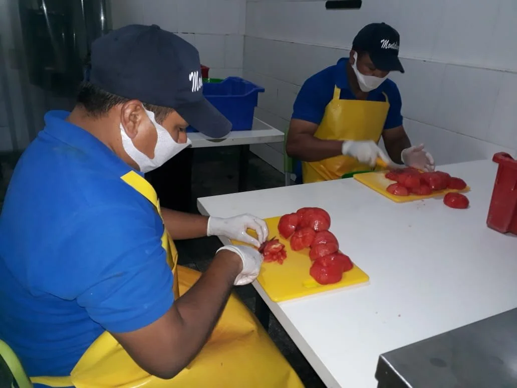 Dos hombres pelando y cortando tomates en una cocina, usando guantes, mascarillas, y gorros.