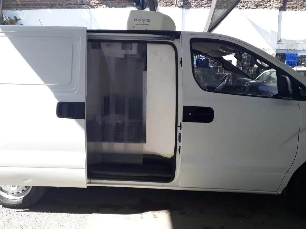 Camión blanco con puerta lateral abierta, mostrando un espacio refrigerado con productos en su interior, estacionado en un área iluminada.