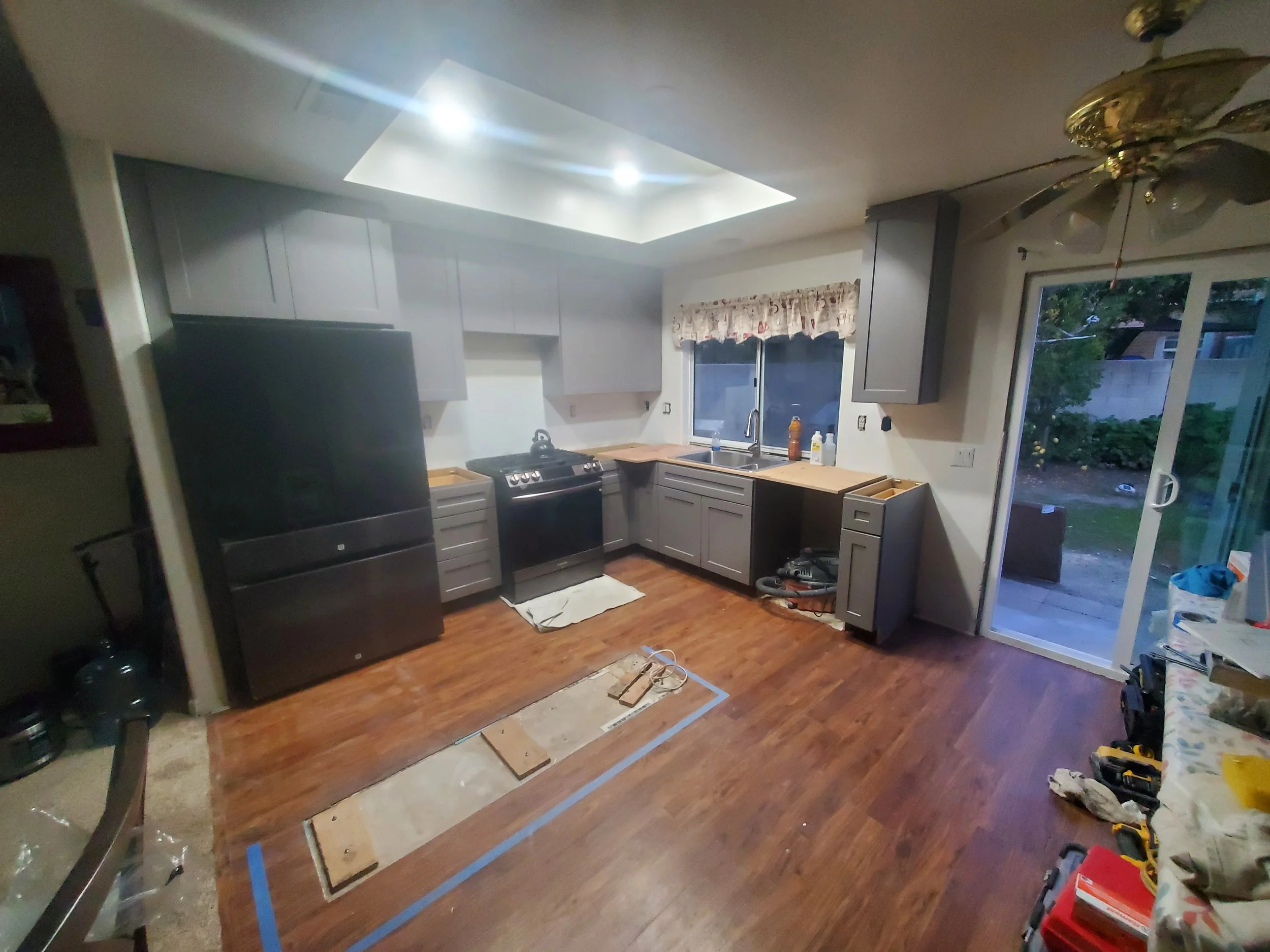Kitchen with gray cabinets, stainless steel appliances, and a wooden countertop. The ceiling has a square recessed light, and a ceiling fan is on the right. A sliding glass door leads outside. There are various tools and materials on the floor, indic