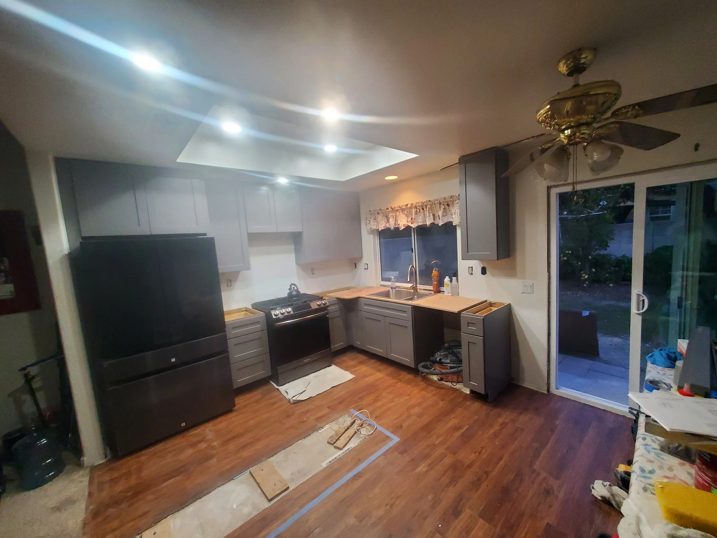 A kitchen with gray cabinets, a black stove, and a sink under a window with a floral valance. An unfinished flooring area with markings and loose painting supplies is in the foreground. There is a sliding glass door to the right leading outside. The 