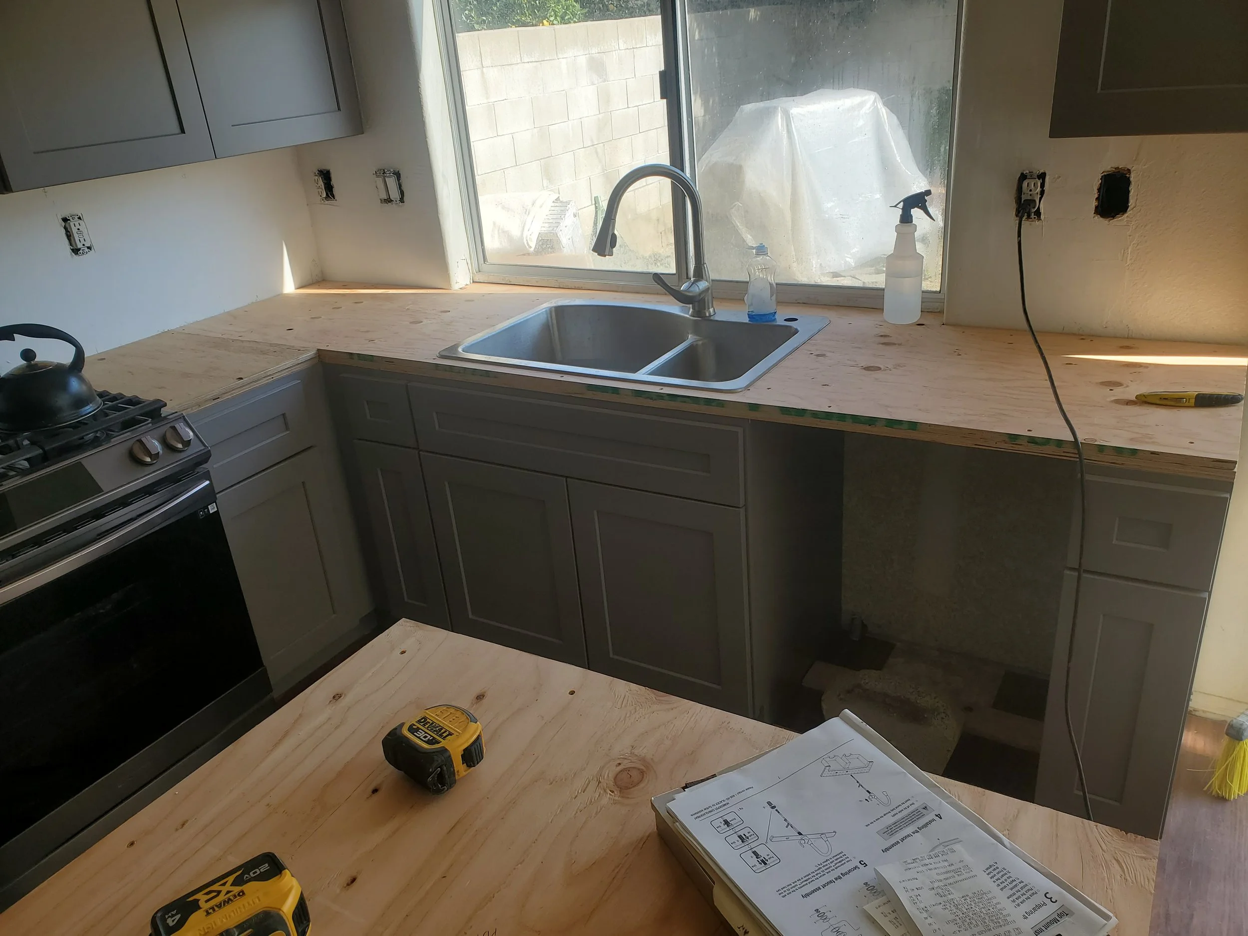A kitchen under renovation with a wooden countertop, a new double sink, and a window above it. Power outlets and switches are missing covers, and tools like a drill and a measuring tape are on the countertop. There is a partially assembled cabinet or