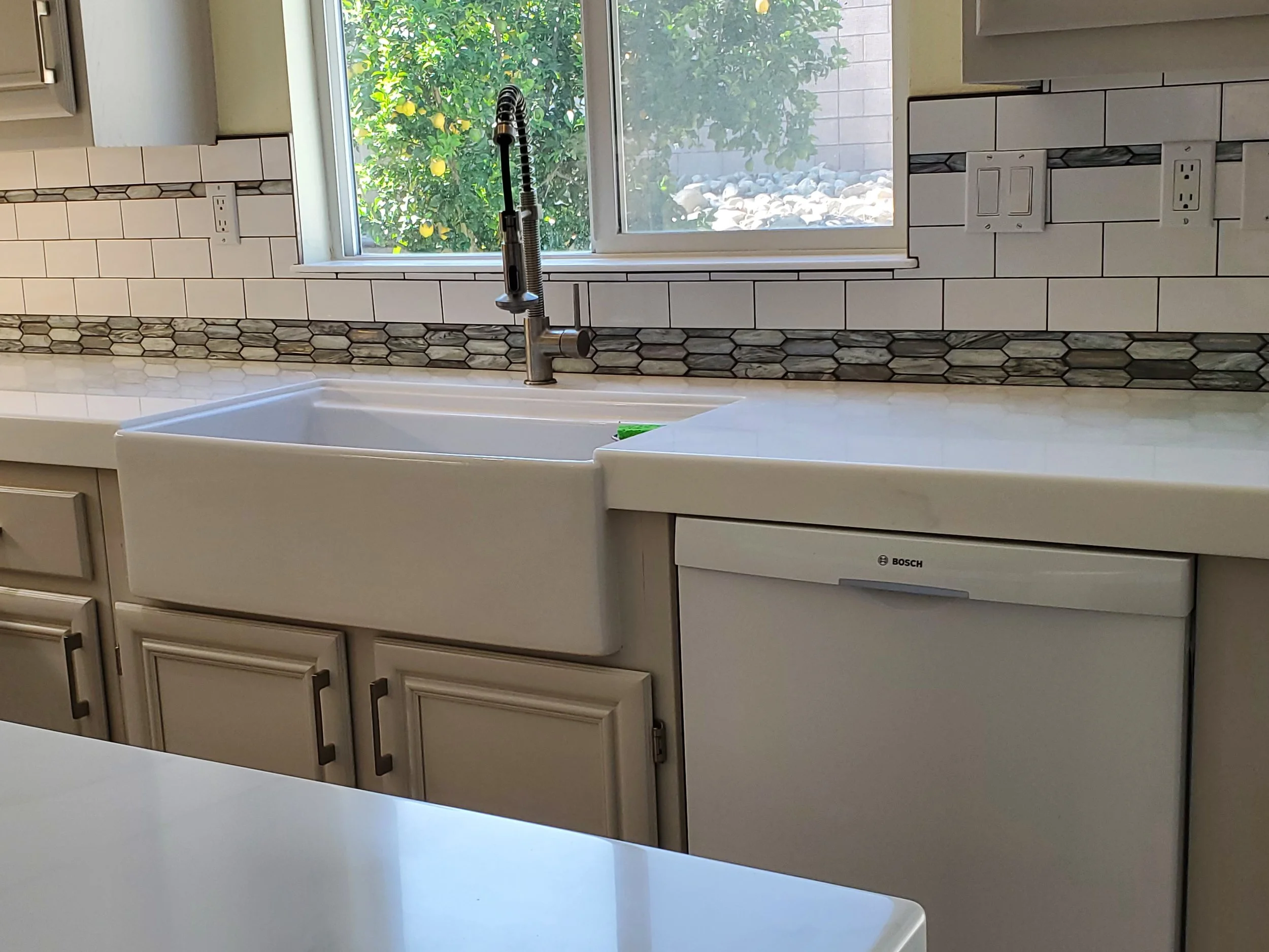 Kitchen sink with a faucet located beneath a window overlooking a lemon tree outside. The backsplash features decorative tiles and electrical outlets.