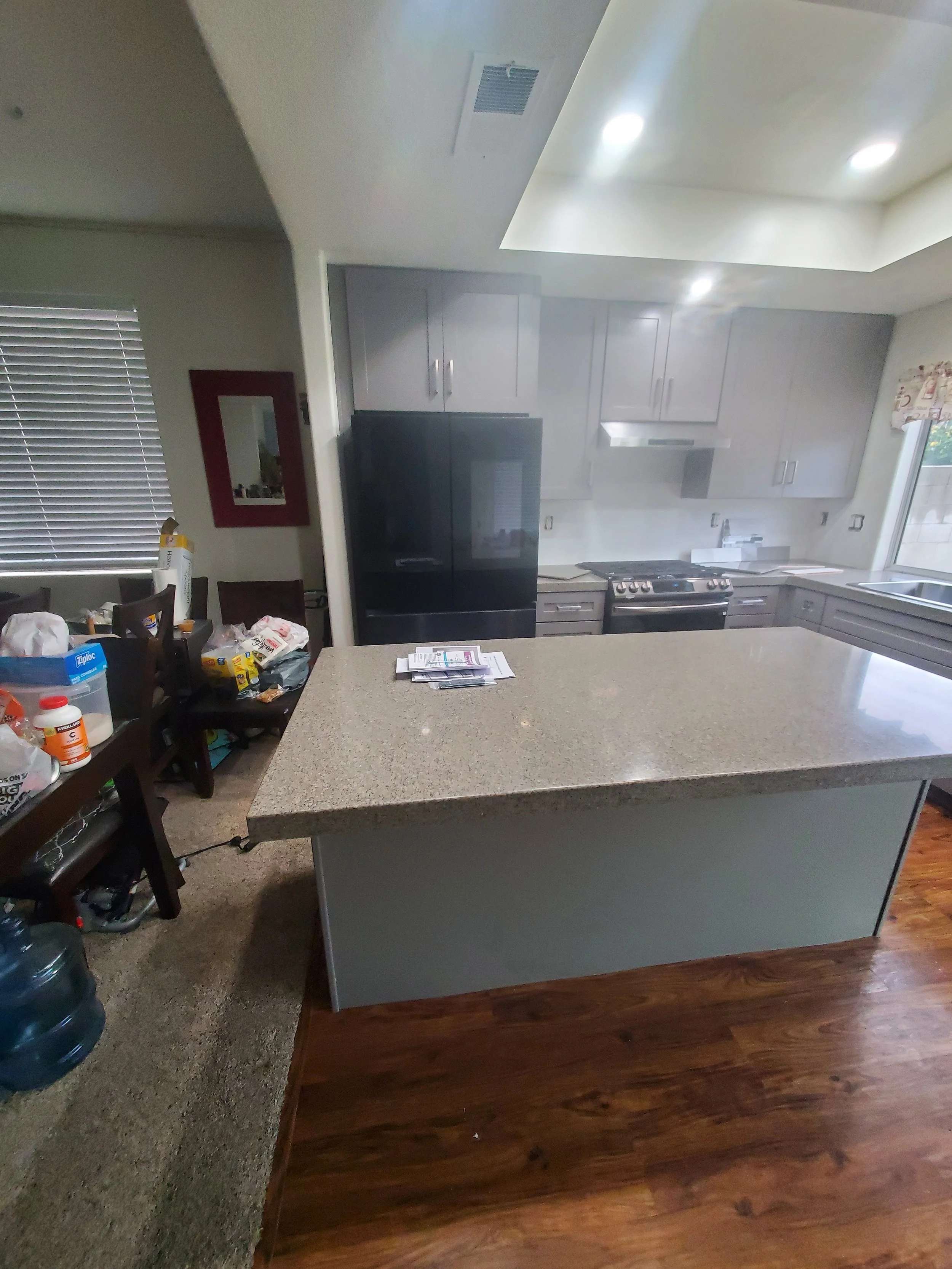 A kitchen with gray cabinets, a stainless steel stove, white countertops, and a stainless steel sink near a window with a valance. There is a large island in the foreground and a dining table with chairs to the left, cluttered with various items.