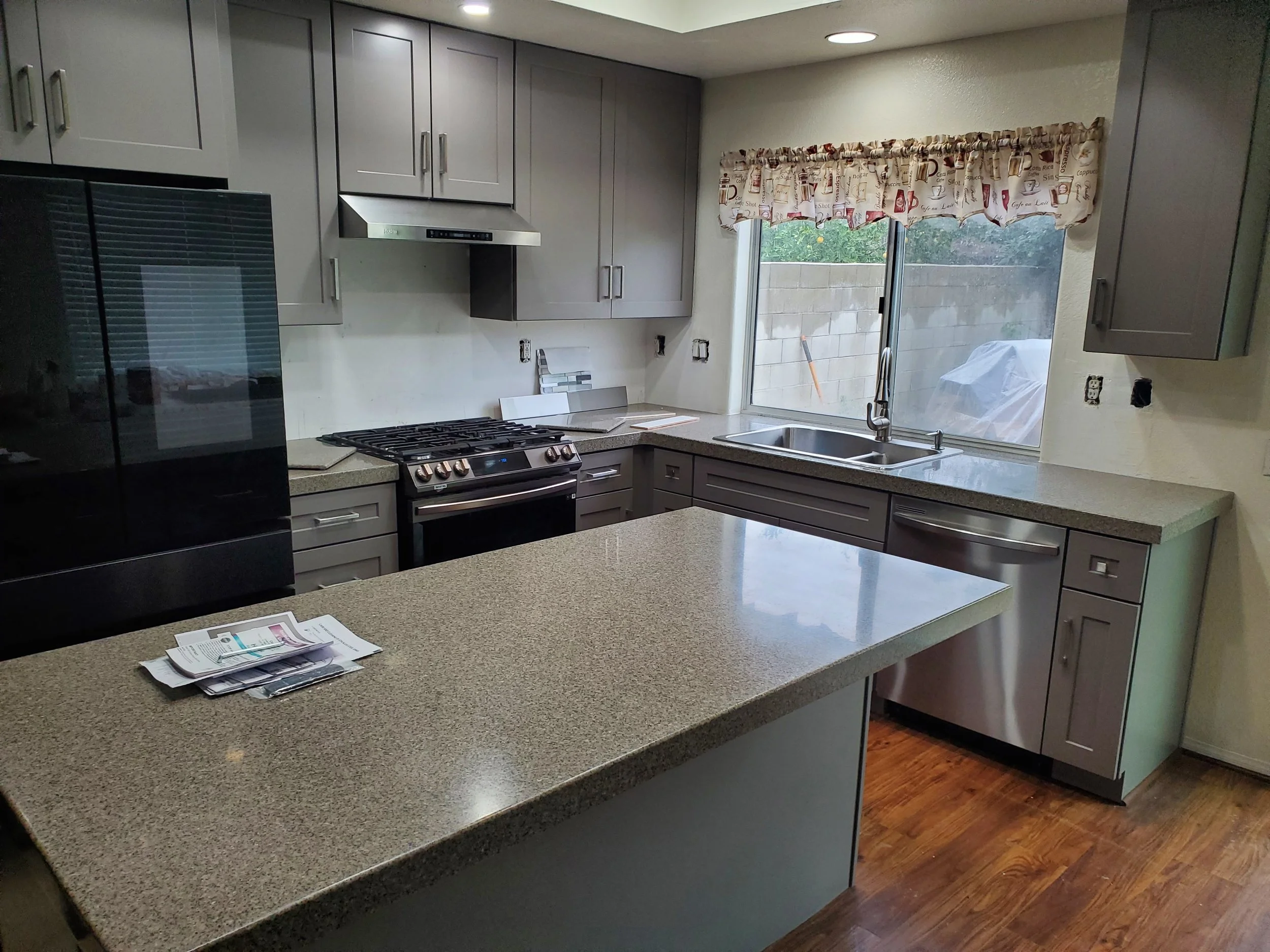 Modern kitchen with gray cabinets and a granite countertop. There is a gas stove with a stainless steel vent hood. A window with a patterned valance overlooks an outdoor fence. The kitchen has wooden floors and some papers on the counter.