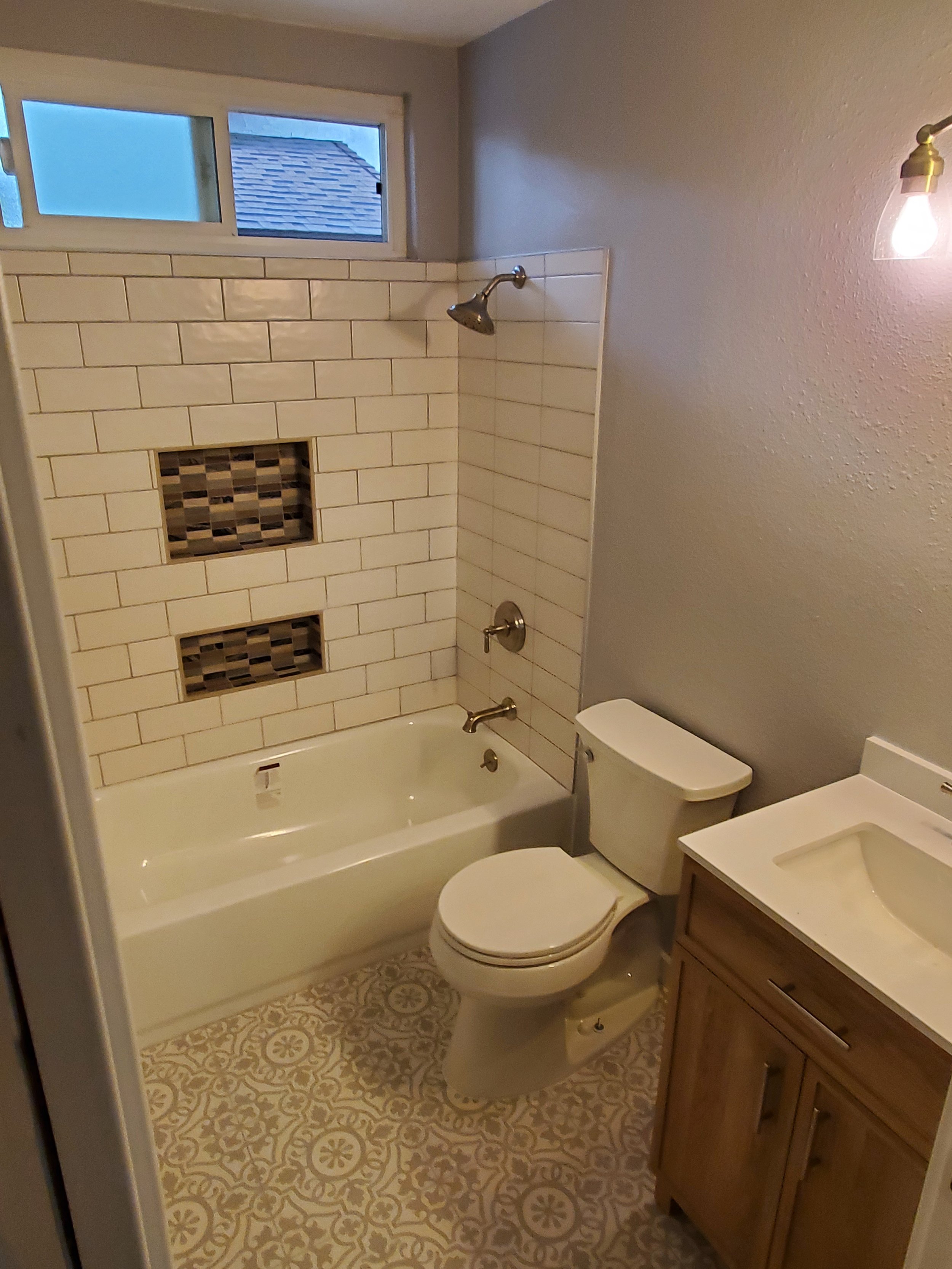A bathroom with a bathtub, showerhead, toilet, and a wooden vanity with a sink. The wall behind the bathtub has white subway tiles with two built-in shelves and a window above for natural light. The floor is patterned with beige and white tiles.