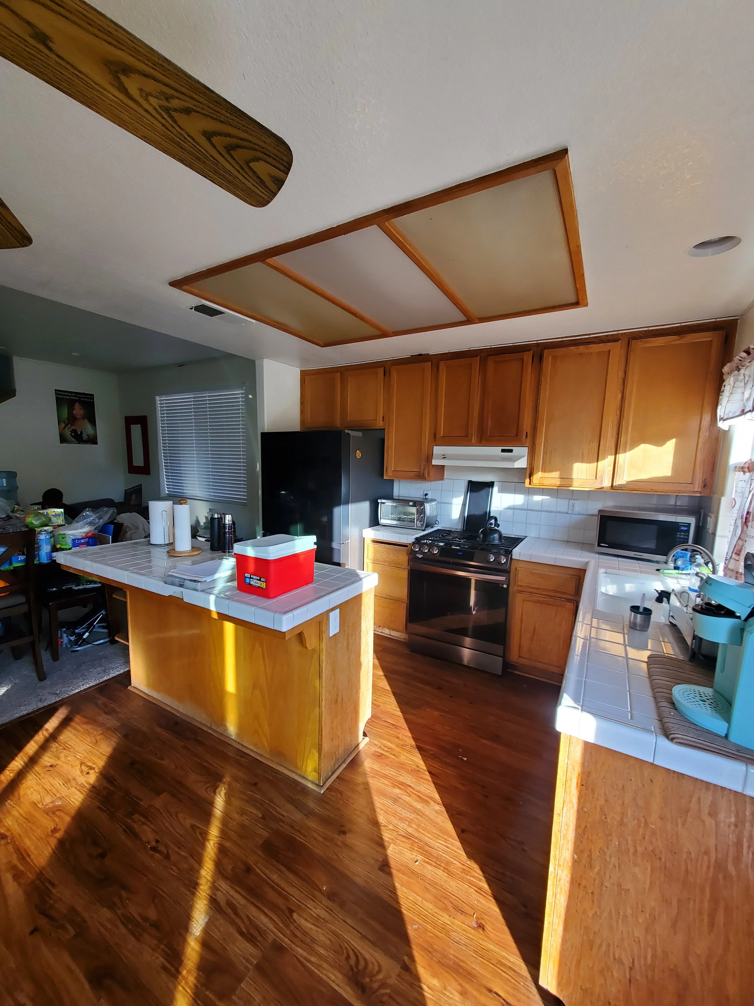 A kitchen with wooden cabinets, a white tiled countertop, a stainless steel stove, a microwave, and a black refrigerator. There is a kitchen island with a white tiled top, holding paper towels, a red cooler, and other items. Sunlight is coming throug