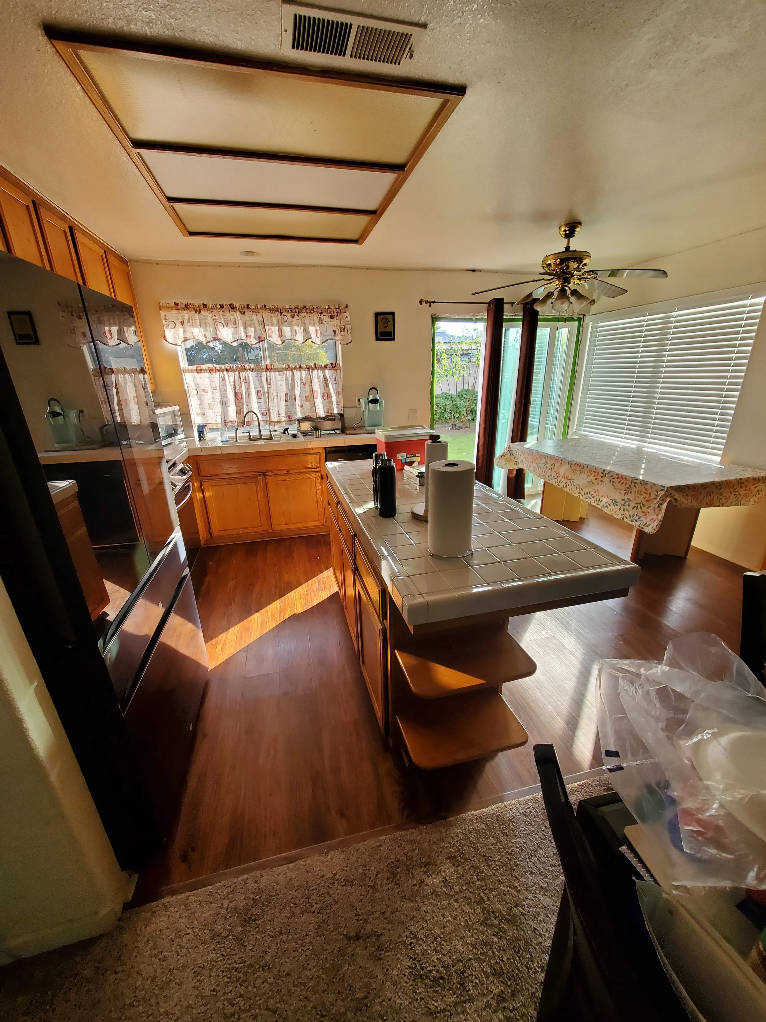 The image shows a kitchen with wooden cabinets, a tiled kitchen island, a ceiling fan, a window with curtains, and sliding glass door leading outside. The counter has various items, including paper towels and a red cooler.