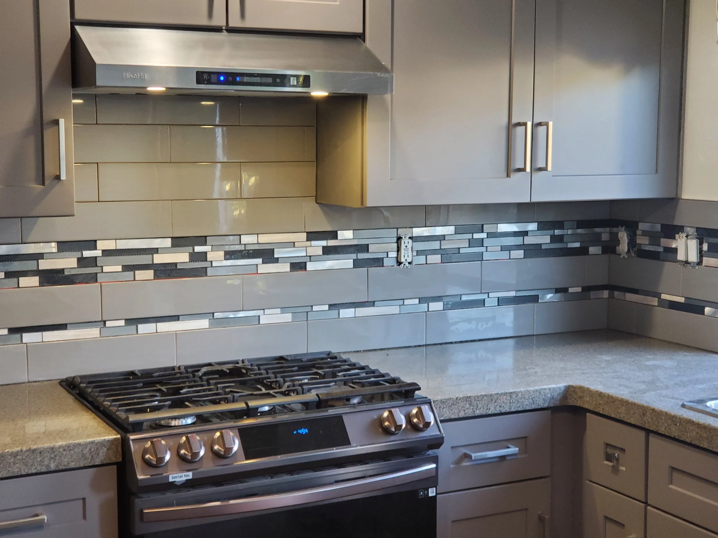 Modern kitchen with gray cabinets, a stainless steel range hood, a black gas stove, and a colorful tile backsplash.