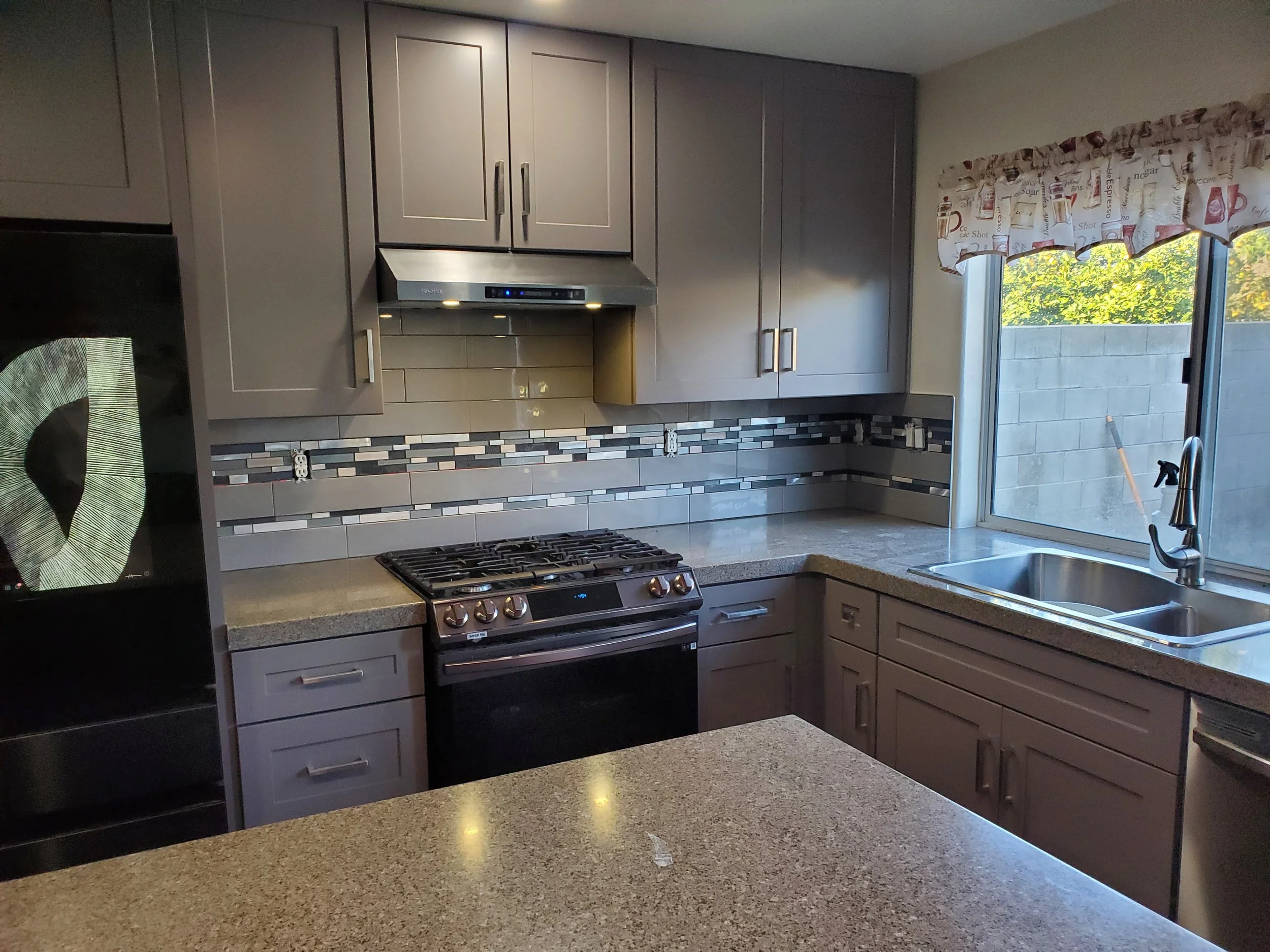 Kitchen with gray cabinets, a gas stove, a granite countertop, and a window with a floral valance. There is a backsplash with horizontal mosaic tiles and electrical outlets along the wall.