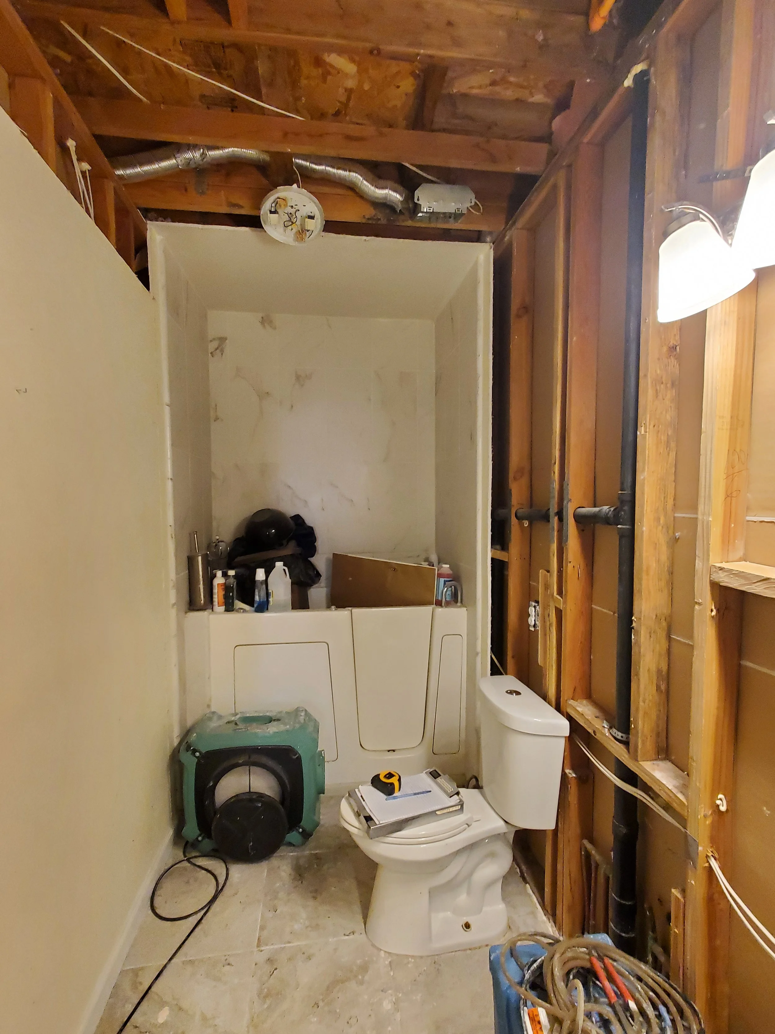 A laundry room under renovation with exposed wooden studs and pipes on the wall, a white toilet, a washing machine, and a dryer. Tools and supplies are scattered around, including a measuring tape and cords, indicating ongoing construction or repair 