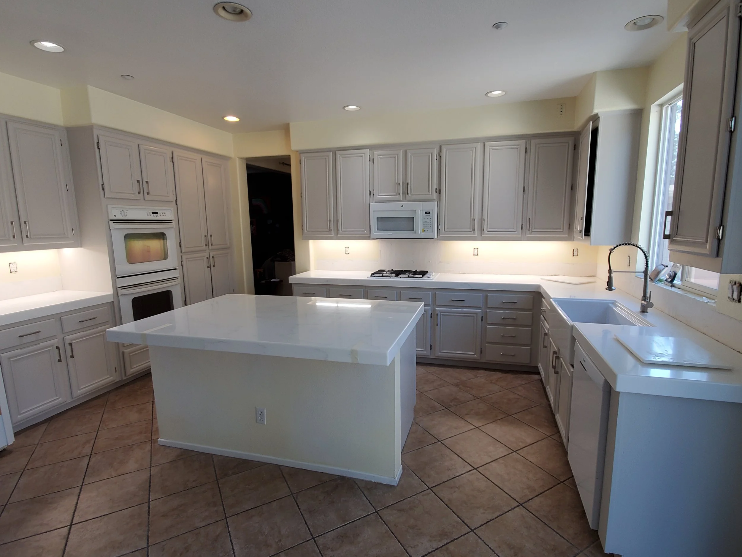 A spacious kitchen with white cabinets, a central island, a white oven and microwave, a sink by the window, and tiled floor.